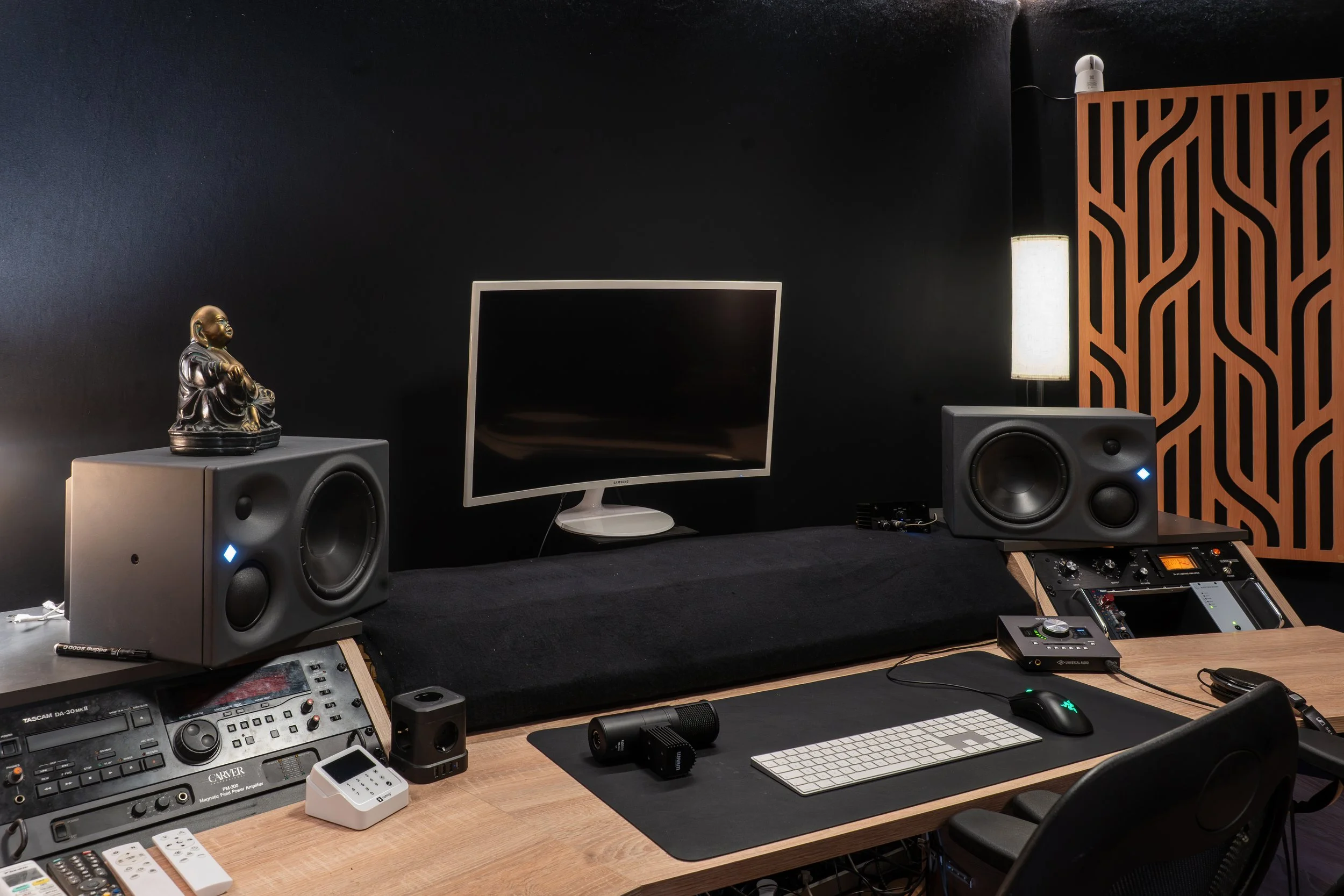 Espace de musique ou studio avec un ordinateur, une console de mixage, des haut-parleurs, et diverses équipements audio sur un bureau en bois. Fond noir avec un panneau en bois décoratif et un éclairage.
