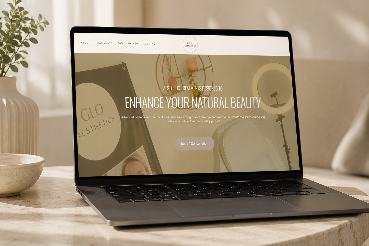 Laptop displaying a website for GLO Aesthetics with the text 'Enhance Your Natural Beauty' and a 'Book A Consultation' button, on a wooden table with a beige bowl and a plant in the background.