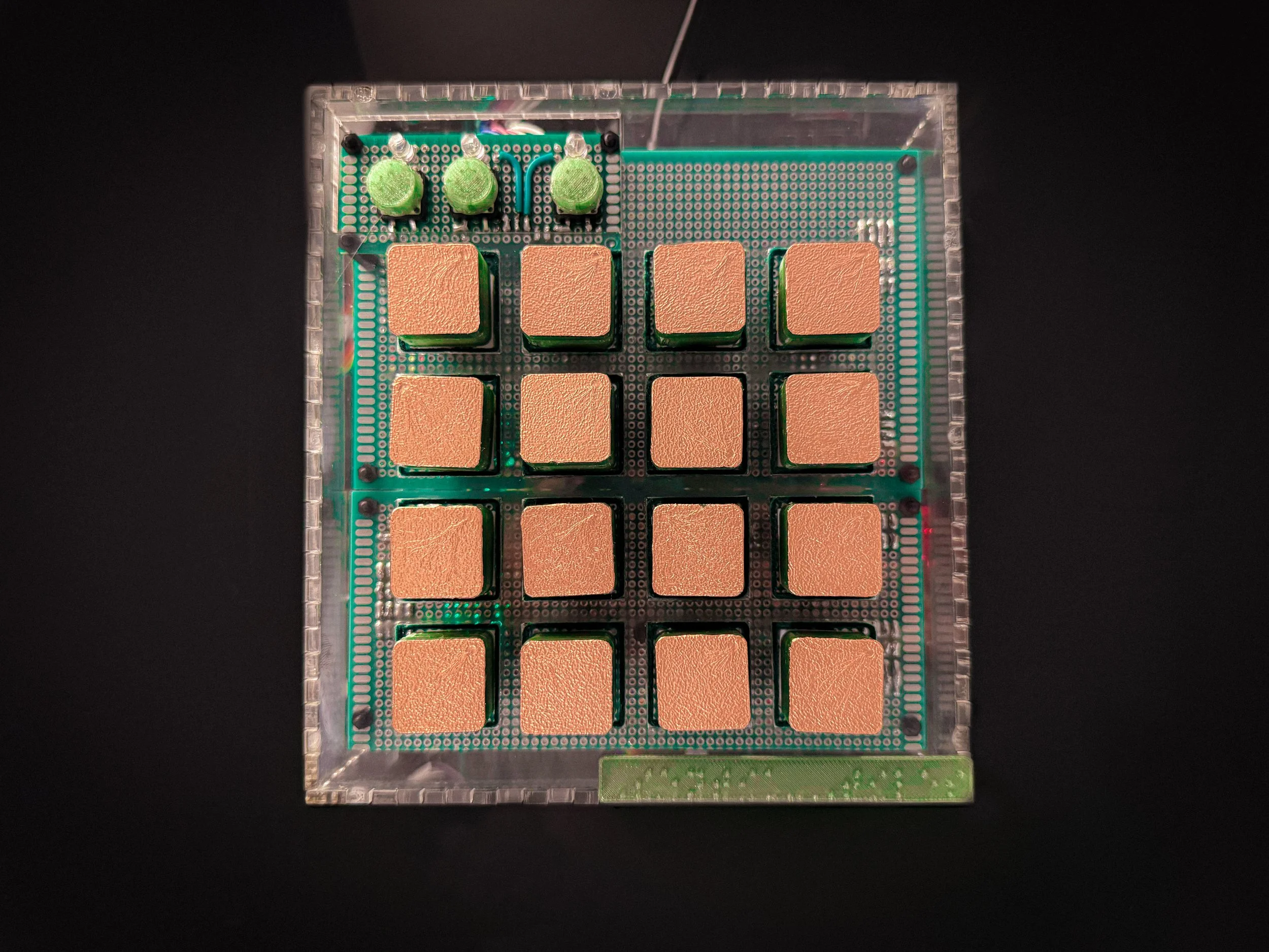 An early prototype of the TILES controller with copper-colored microprocessors and green components, enclosed in a clear plastic case, with a black background.