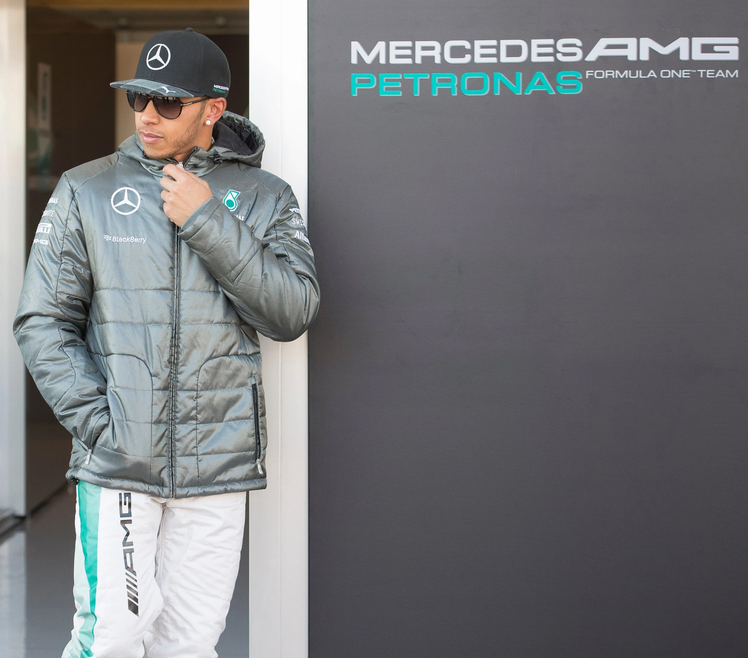 A male athlete dressed in Mercedes AMG Petronas Formula One Team apparel standing against a wall with the team logo, wearing sunglasses, a cap, and a puffy jacket