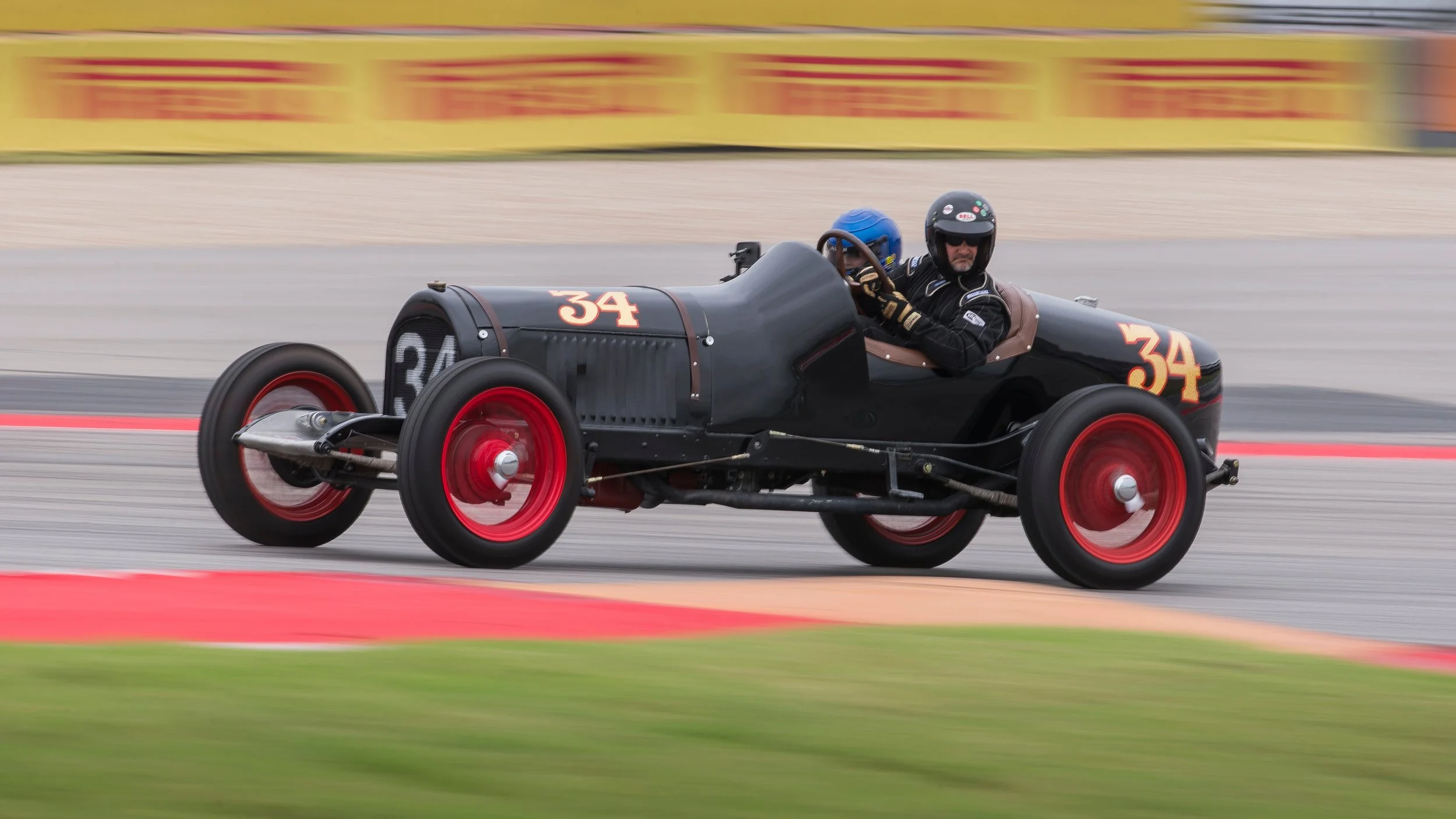Vintage black race car with red wheels numbered 34 speeds along a race track, driven by a driver wearing a black helmet and racing suit, with a blue helmeted passenger in the backseat.