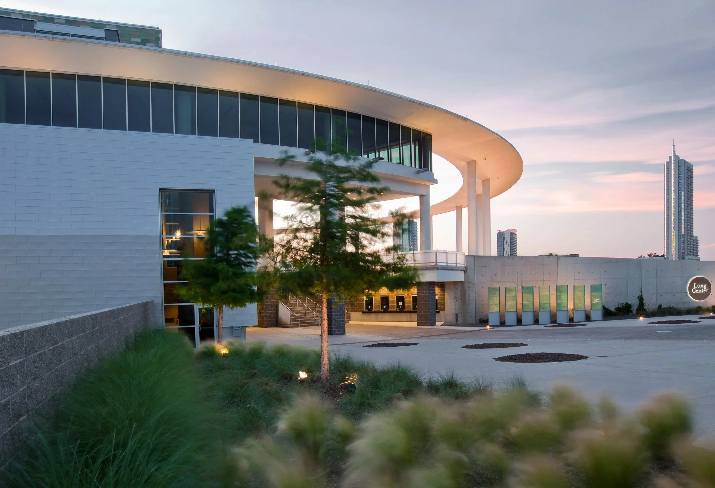 Modern building with large glass windows and a curved roof, illuminated in the early evening with city skyscrapers in the background, landscaped with small trees and grass.