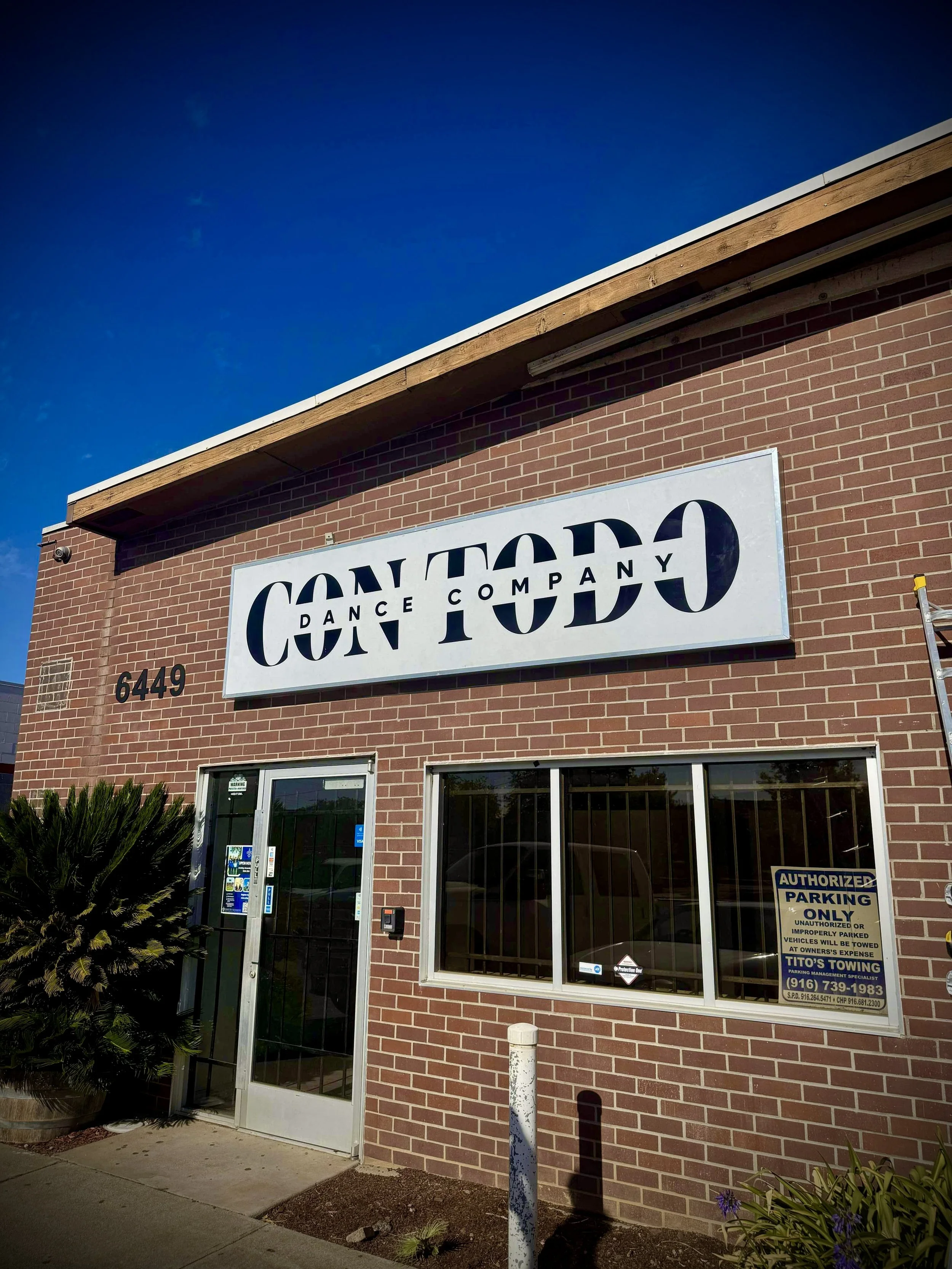 A brick building with a sign that reads 'Con Teco Dance Company' and a parking sign outside. Clear blue sky in the background.