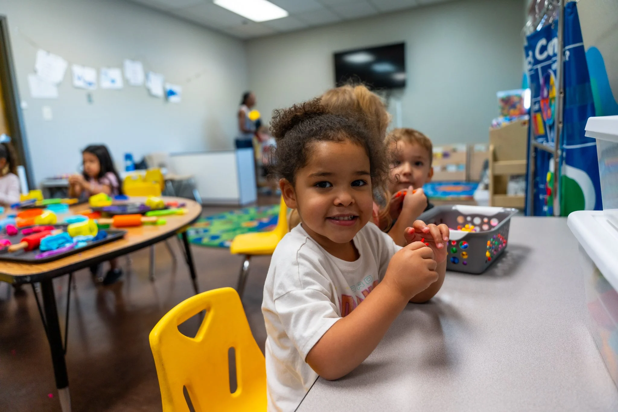 Bright-eyed young girl with curly hair smiling at the camera in a classroom, surrounded by other children and colorful art supplies.