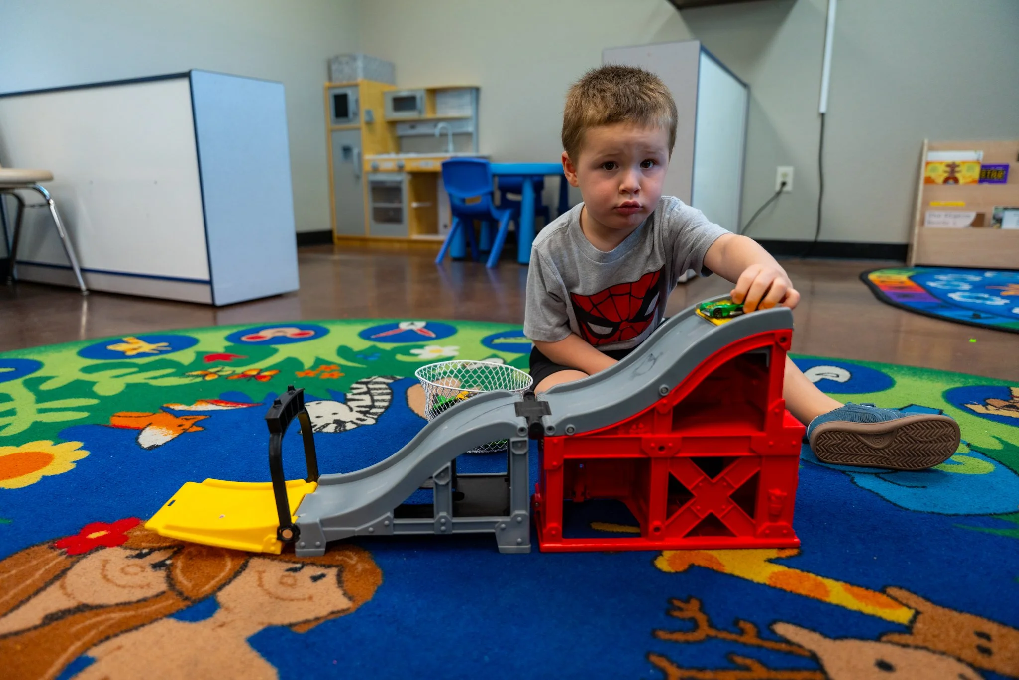 A young boy with brown hair sitting on a colorful play carpet in a room, playing with a toy parking garage and car. The room has children's furniture and a bookshelf in the background.