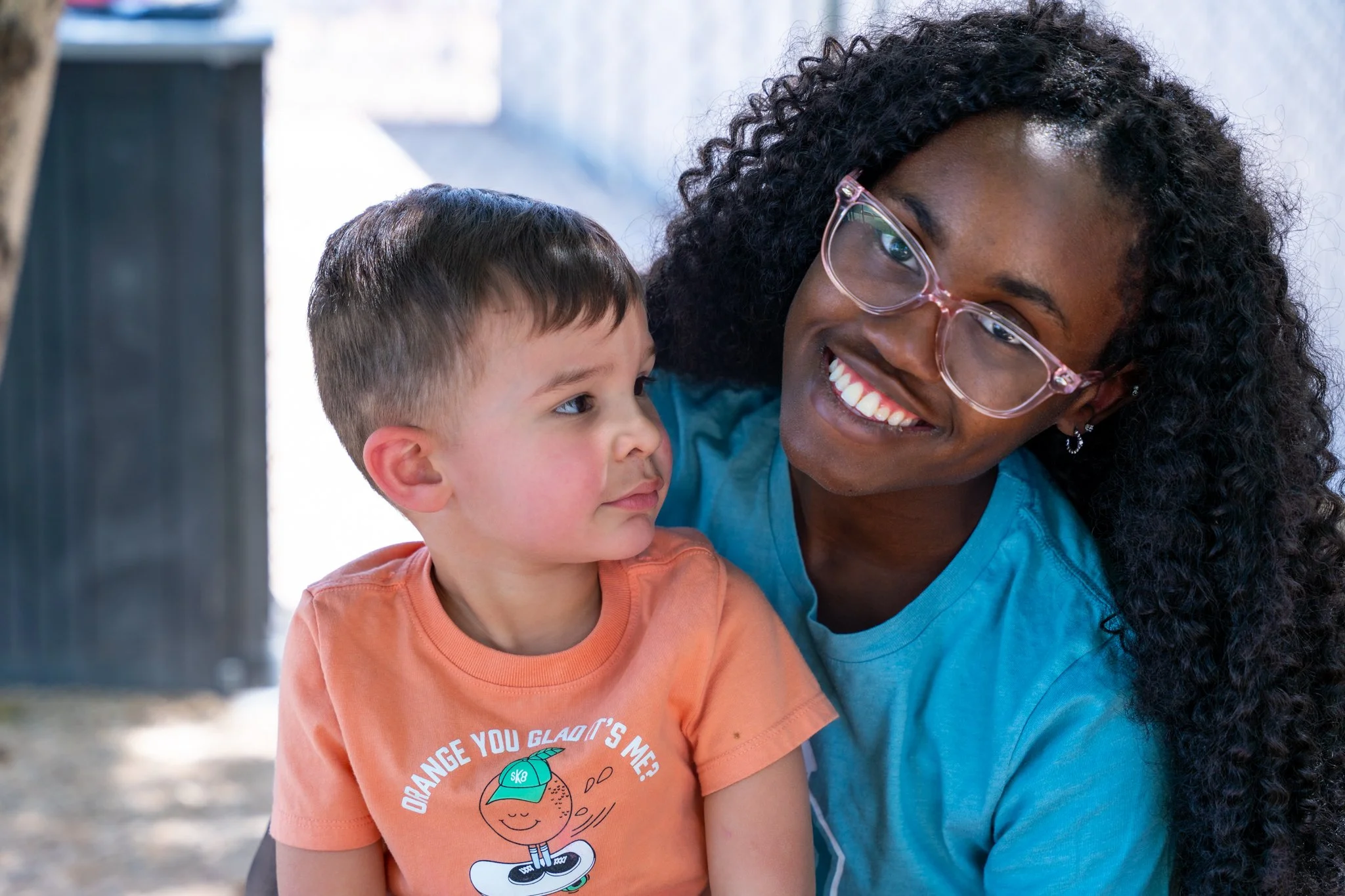 A smiling woman with curly hair and glasses is holding a young boy with short brown hair and a peach-colored T-shirt. They are outdoors with a blurred background.