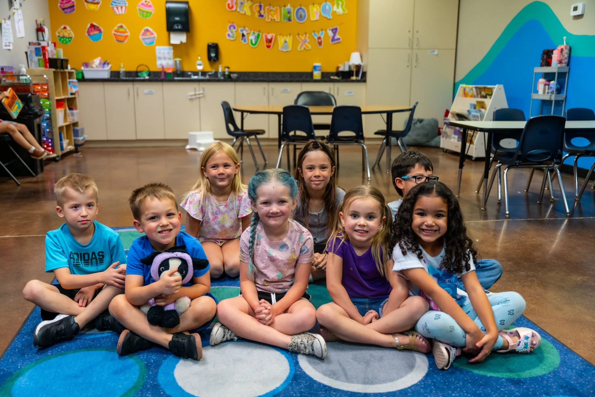 Group of young children sitting on a colorful rug in a classroom, smiling at the camera. The classroom has yellow and blue walls with colorful decorations and children's books and toys on shelves.