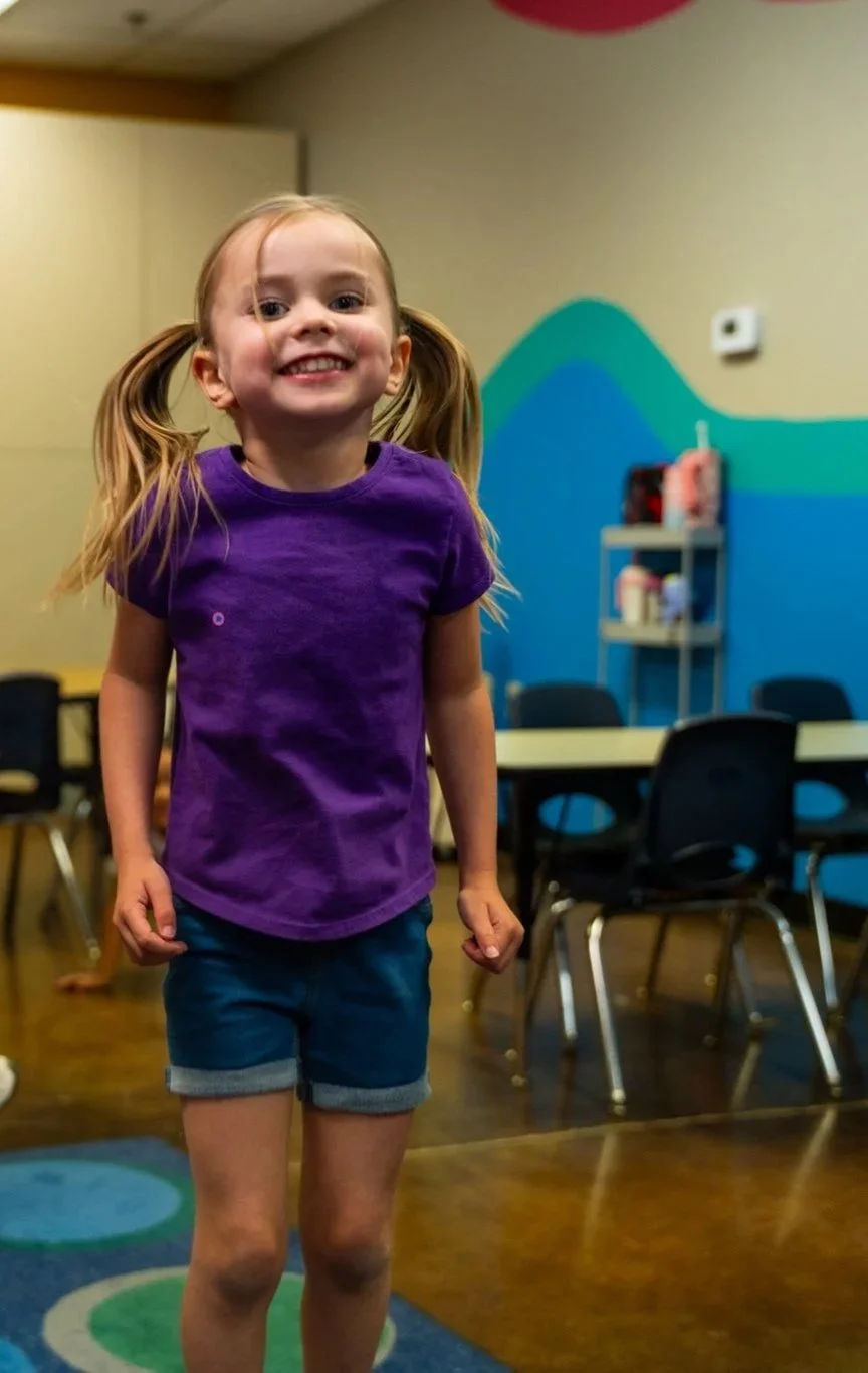 Young girl with pigtails wearing a purple shirt and denim shorts, smiling cheerfully indoors with colorful wall art and chairs in the background.