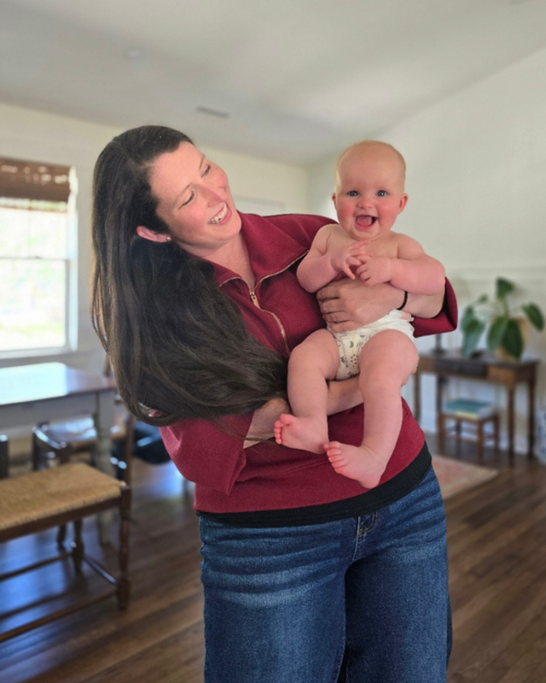 A woman holding a smiling, happy baby in a living room with wooden floors and furniture, large windows, and a potted plant in the background.