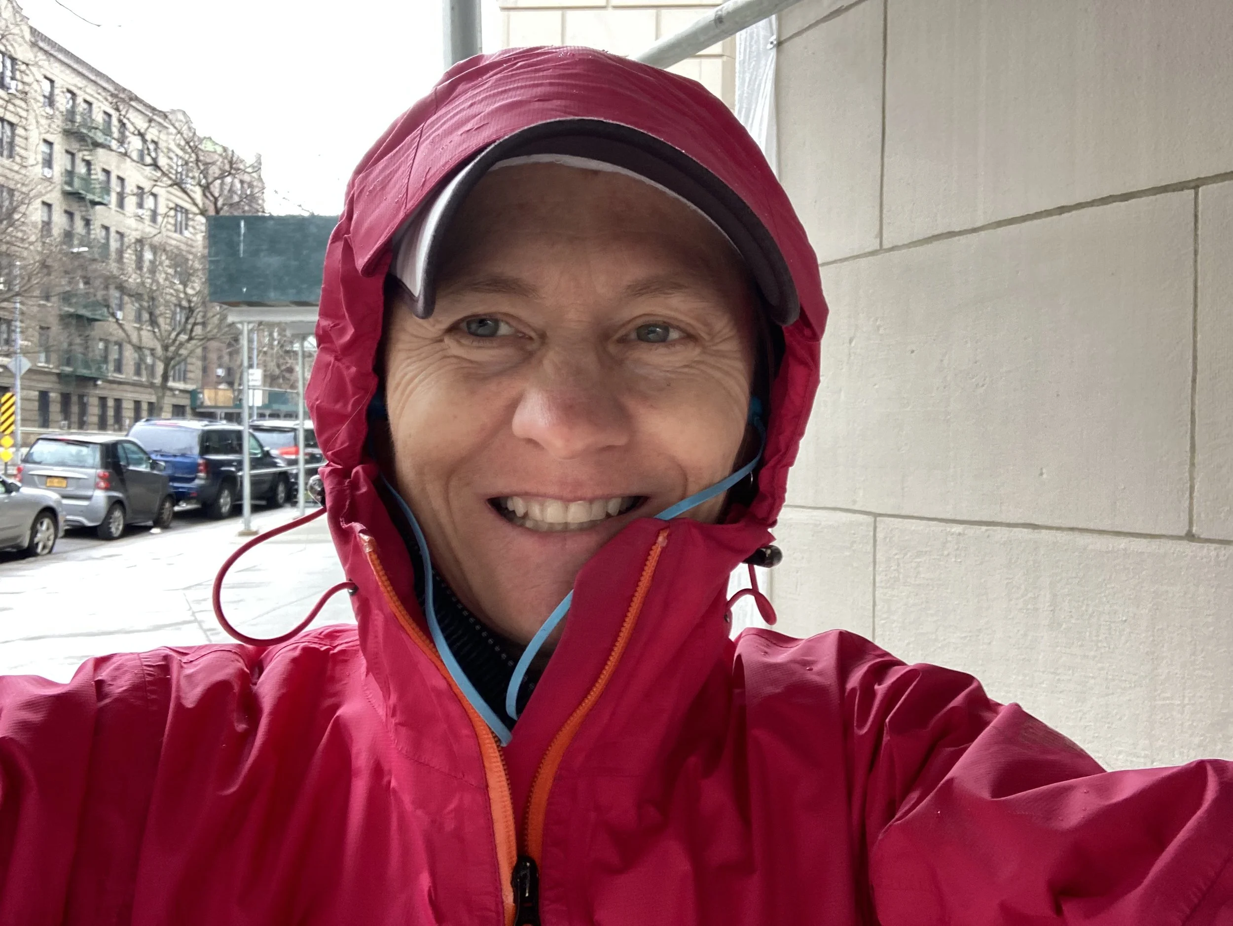 Close-up of a smiling woman in a red jacket and matching hood, wearing a baseball cap and sunglasses, outdoors in an urban setting with parked cars and apartment buildings in the background.