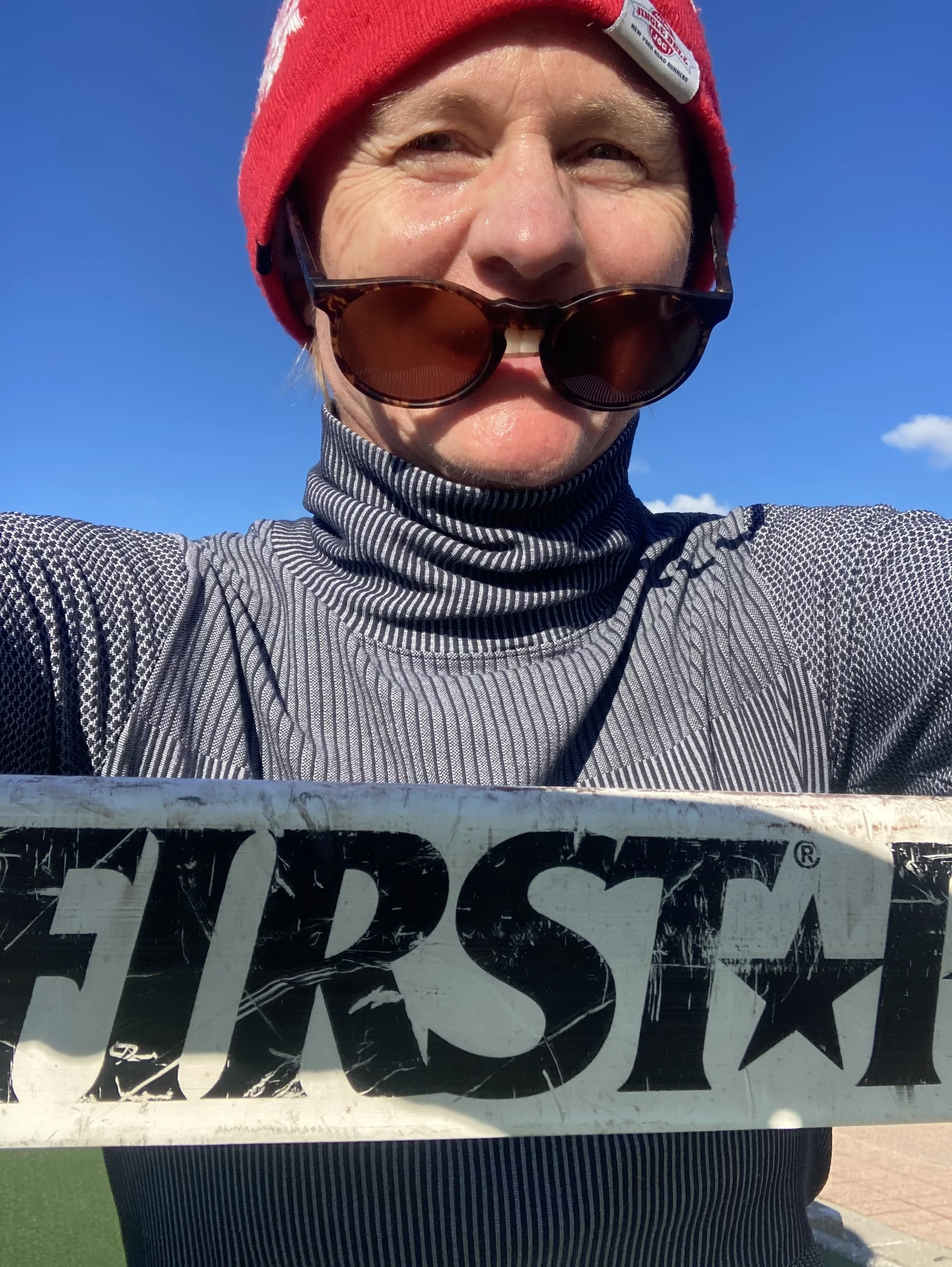 Person wearing a red knit cap, sunglasses, and a gray textured long-sleeve shirt, holding a black object with white and black text in front of a clear blue sky.