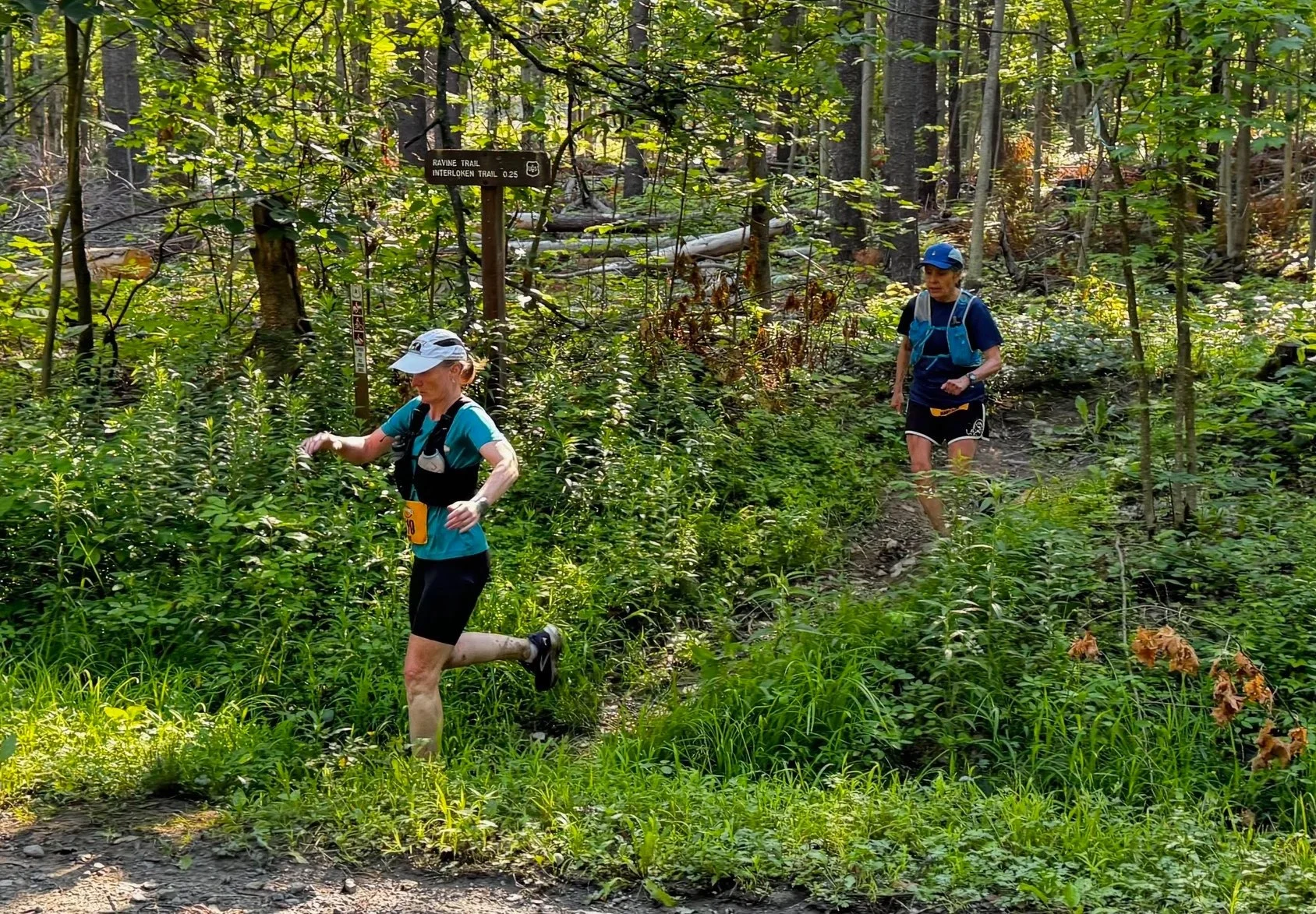 Two competitors trail running through a lush green forest with a trail sign visible in the background.