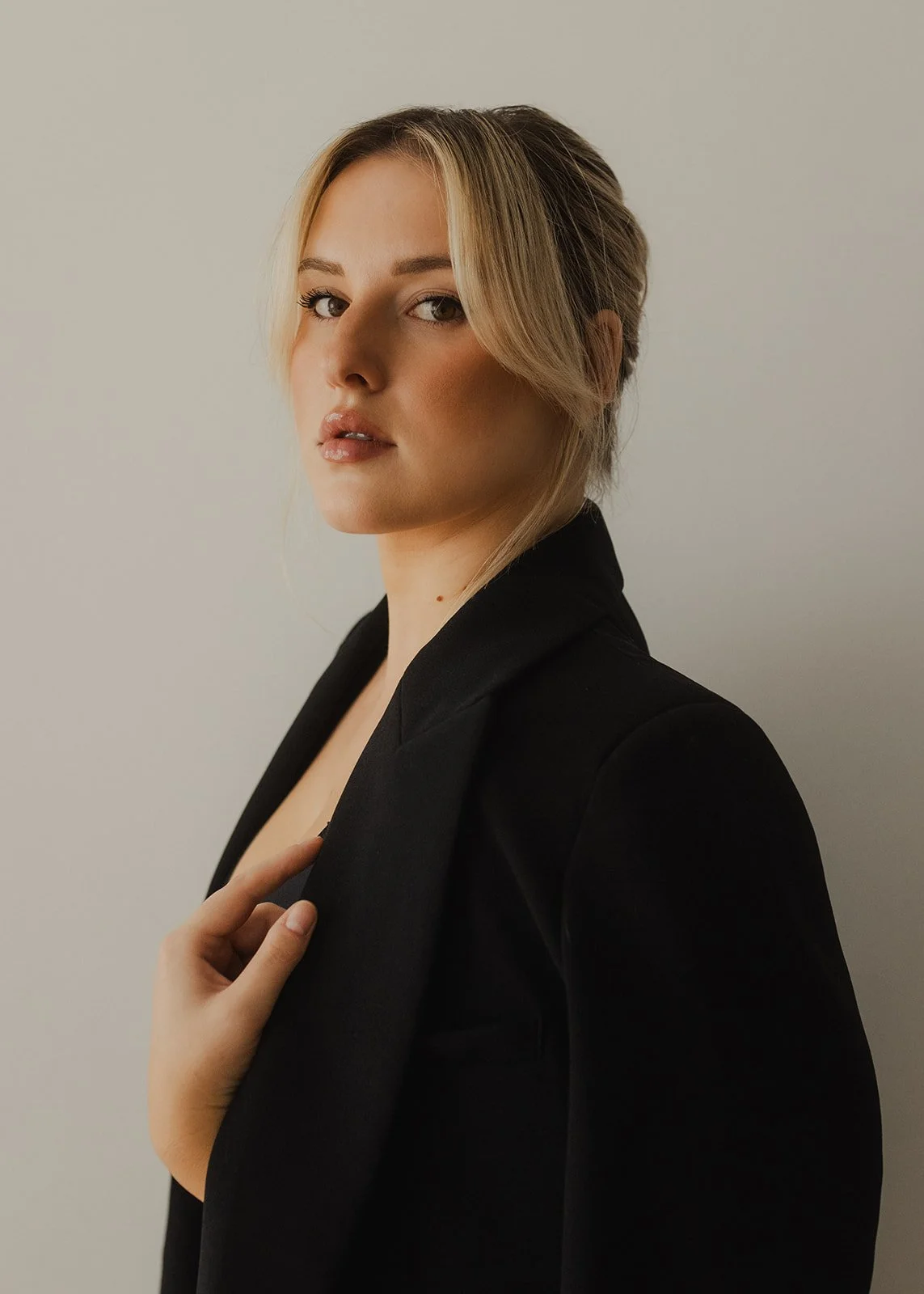 Young woman with blonde hair in a loose updo, wearing a black blazer, standing against a plain light-colored background.