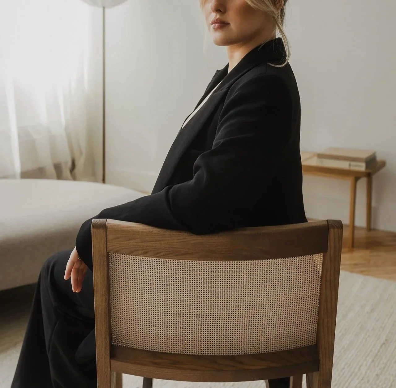 A woman in a black blazer sitting on a wooden chair in a minimalistic room with a bed, a side table, and books, with her hands resting on her lap.