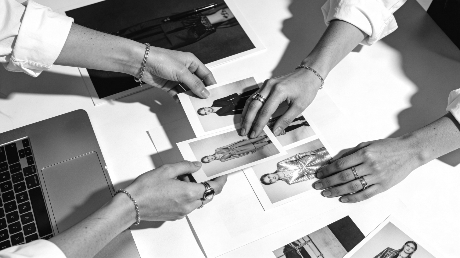 Two people are looking at photographs of models in fashion outfits on a table, with a laptop nearby and wearing bracelets and rings.