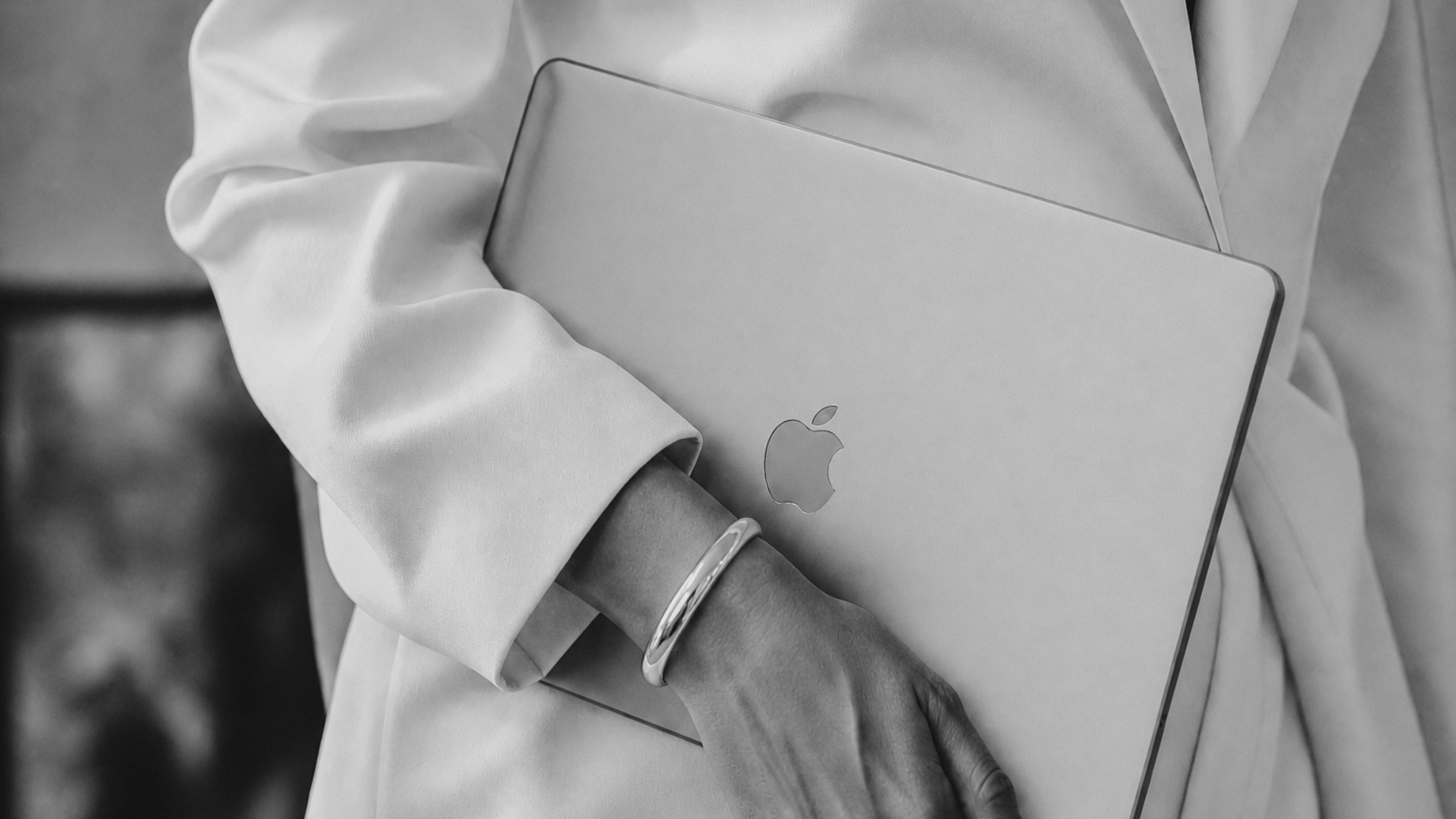 Person holding a closed silver MacBook laptop, wearing a light-colored jacket with rolled-up sleeves, a silver bracelet, and a watch.