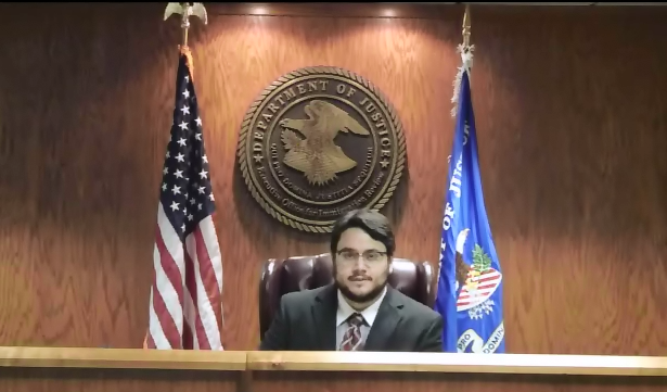 A young José Pitti with glasses and a beard sitting at an immigration judge's bench, as part of a DOJ training video, with the Department of Justice seal on the wall behind him, and two flags on either side.