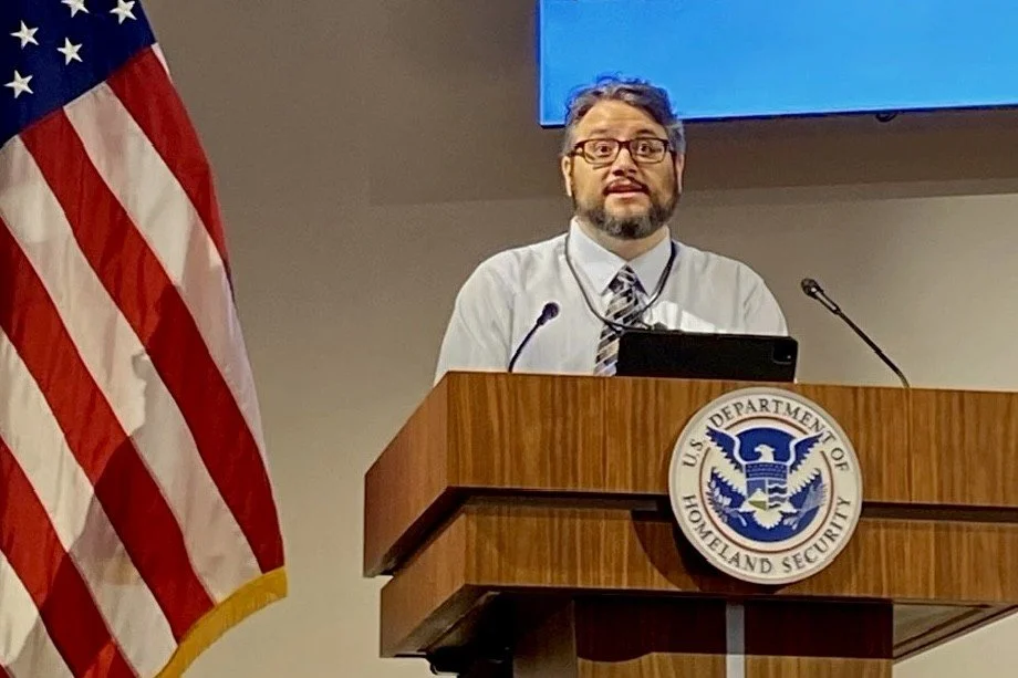 José Pitti standing behind a wooden podium with the U.S. Department of Homeland Security seal, speaking into microphones about civil rights investigations. An American flag is to his left.