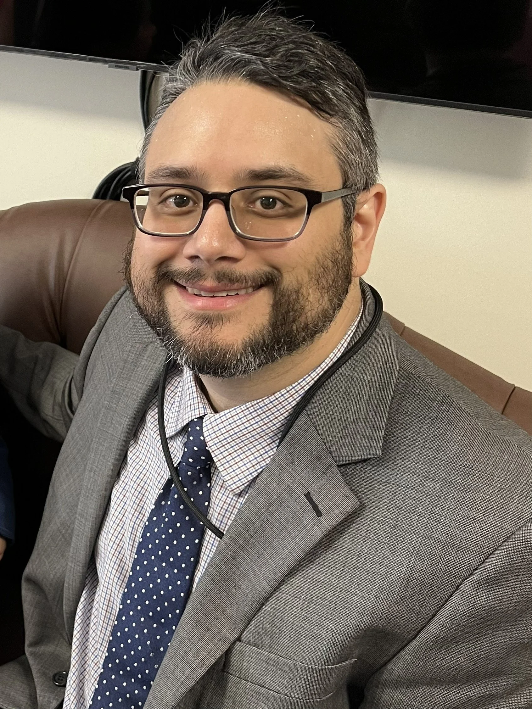 José Pitti smiling, wearing glasses, a gray suit, and a polka dot tie, sitting on a leather couch.