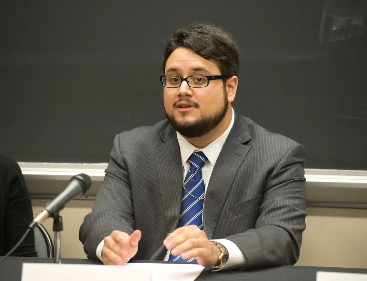 A young José Pitti in a gray suit, white shirt, and striped blue tie is seated at a table speaking, with a black chalkboard in the background. He has glasses, a beard, and is gesturing with his hands while speaking.