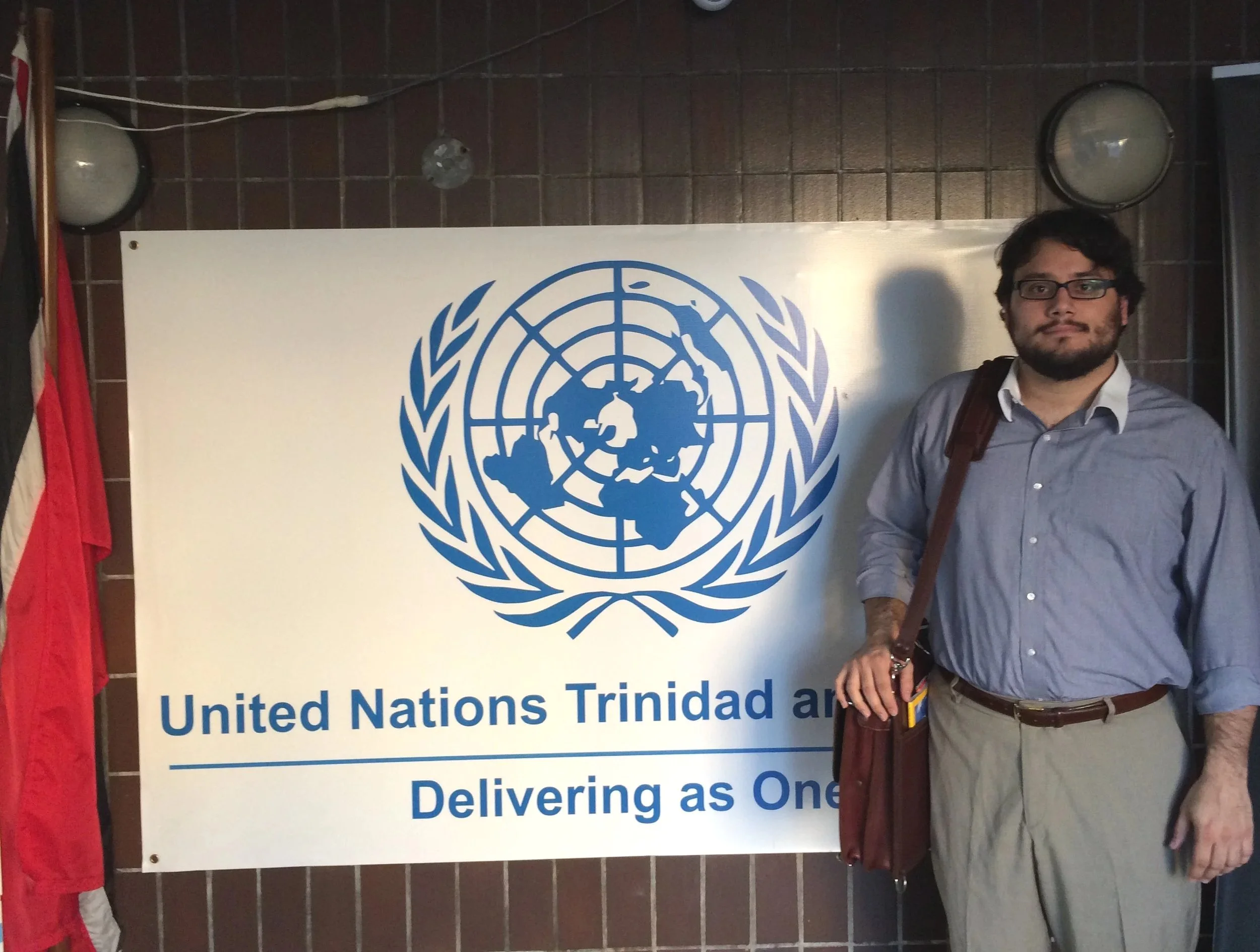 José Pitti standing next to a United Nations Trinidad and Tobago sign, holding a bag, in front of a dark tiled wall with two wall-mounted lights and a red flag.