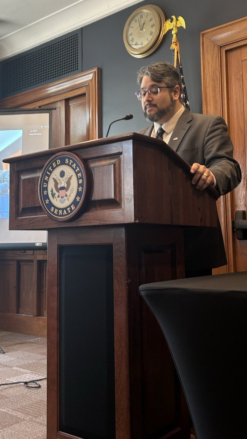 José Pitti in a suit and glasses standing at a wooden podium with the United States Senate seal, speaking into a microphone. There is a clock and an American flag behind him in a formal room in a U.S. Senate Building.