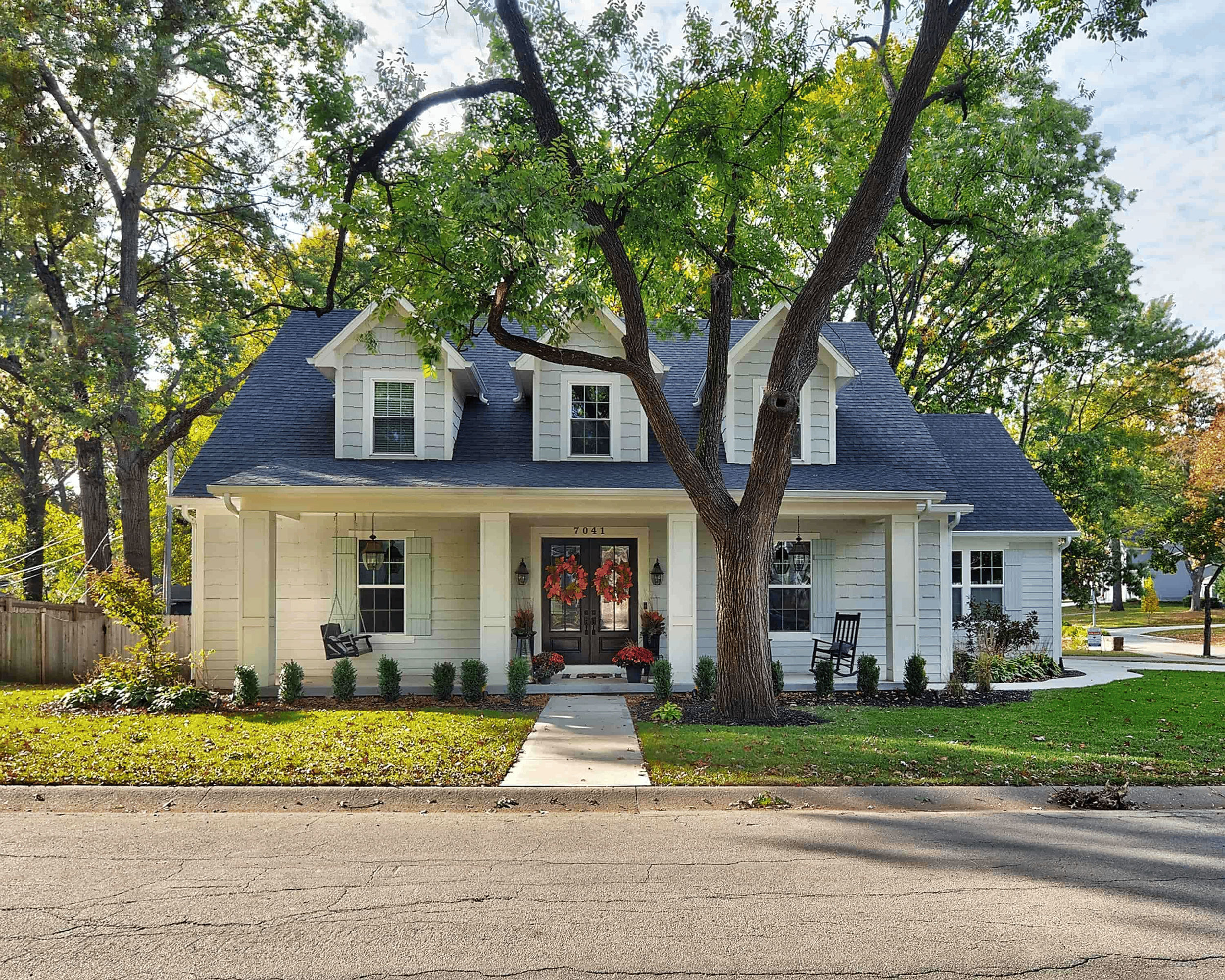 Front view of a house with a pitched roof, three dormer windows, and a welcoming porch decorated with flower pots and outdoor chairs. The house is surrounded by lush green trees and a well-maintained front yard, with a sidewalk and street in the foreground.