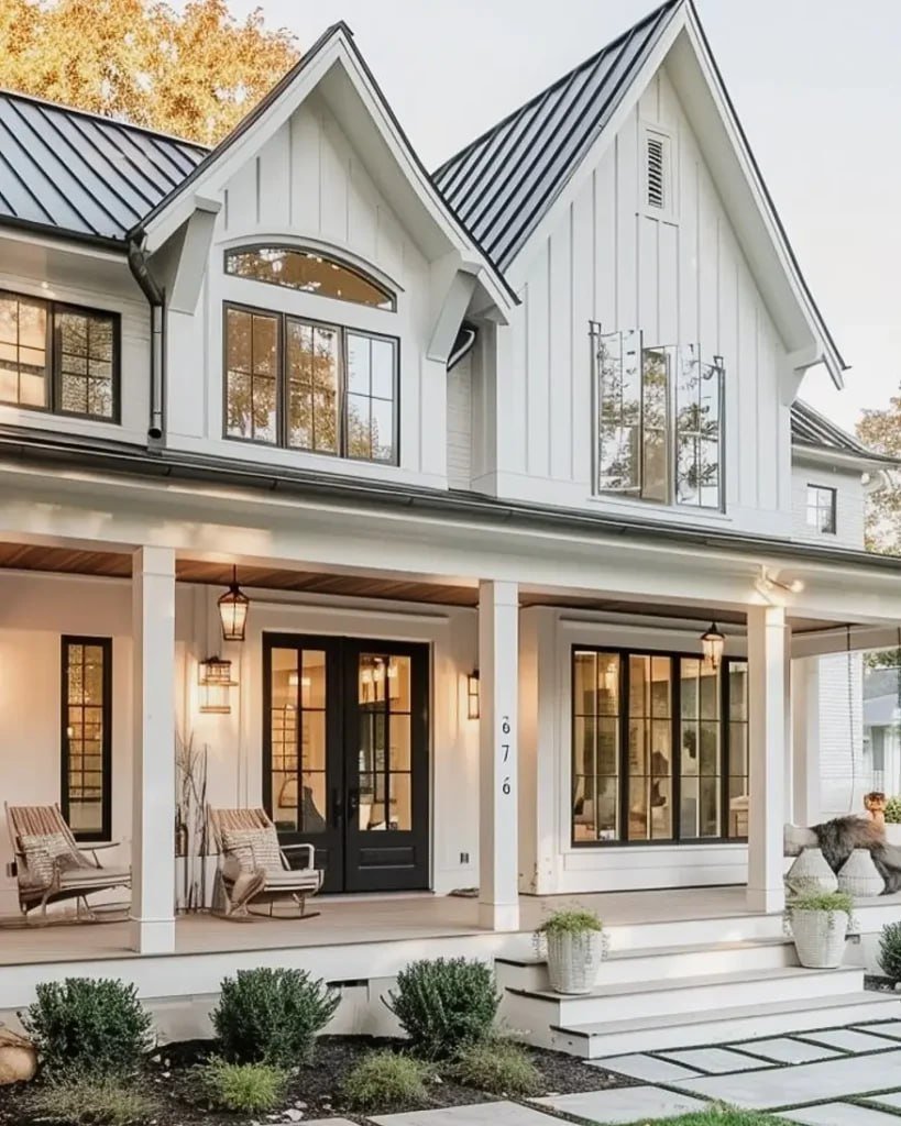 A two-story modern farmhouse with white siding, black window frames, and a metal roof, featuring a front porch with seating and potted plants.