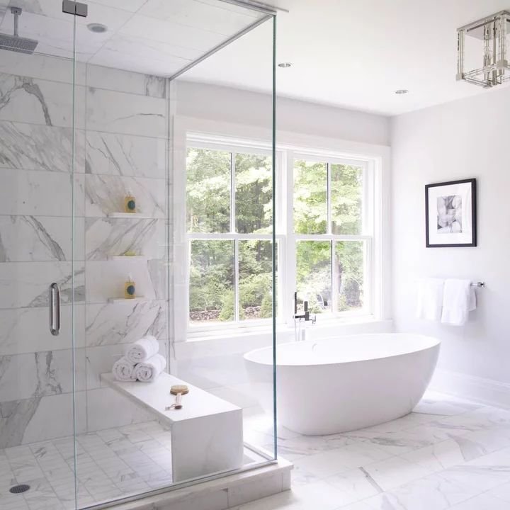 Modern bathroom with freestanding bathtub in front of large window, marble flooring and tiles, glass shower with built-in shelves, white walls, and minimalistic decor.