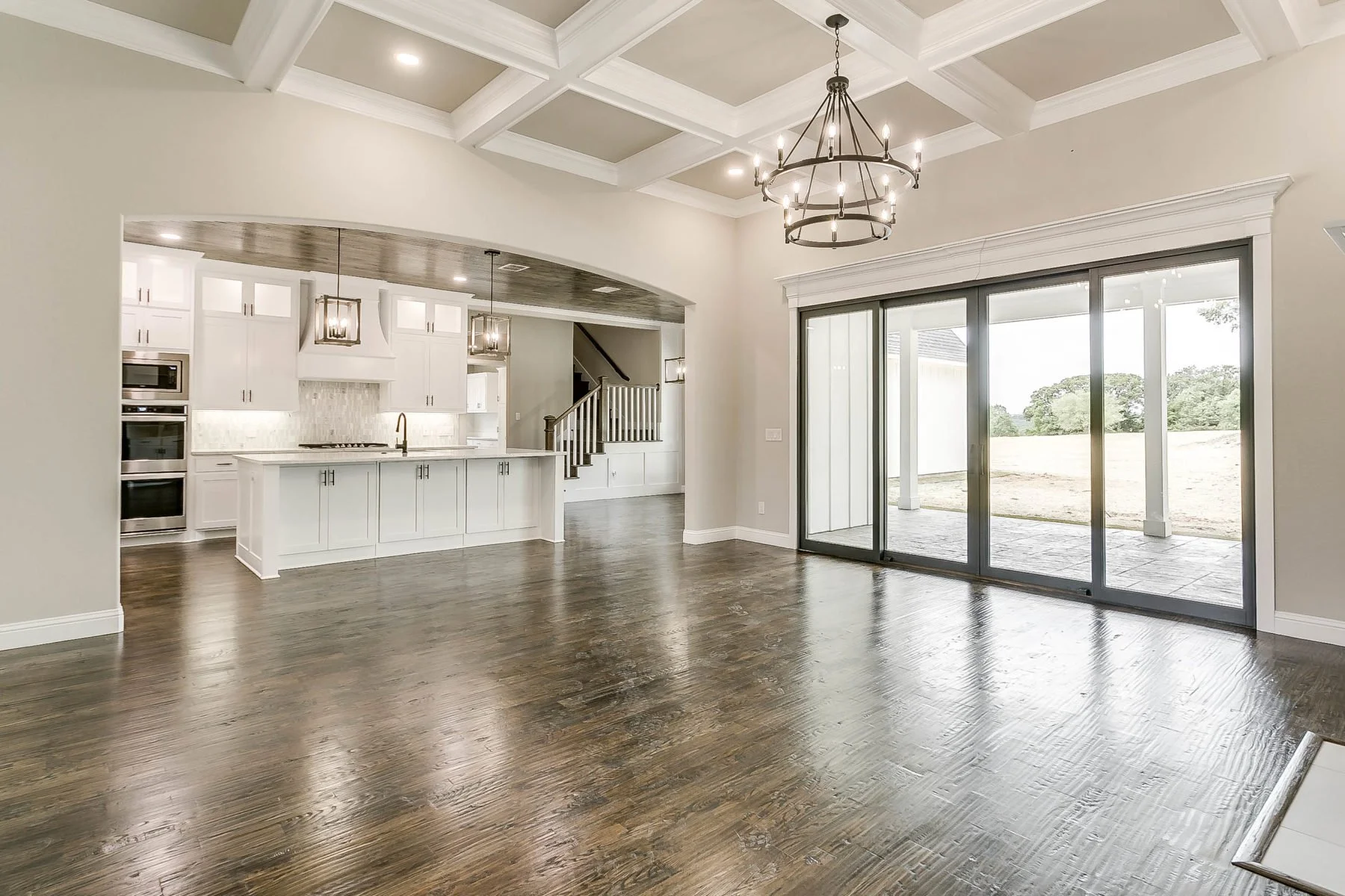 Open-concept living space with hardwood floors, white kitchen cabinets and a kitchen island, large sliding glass doors leading to an outdoor patio, and a decorative chandelier hanging from a coffered ceiling.