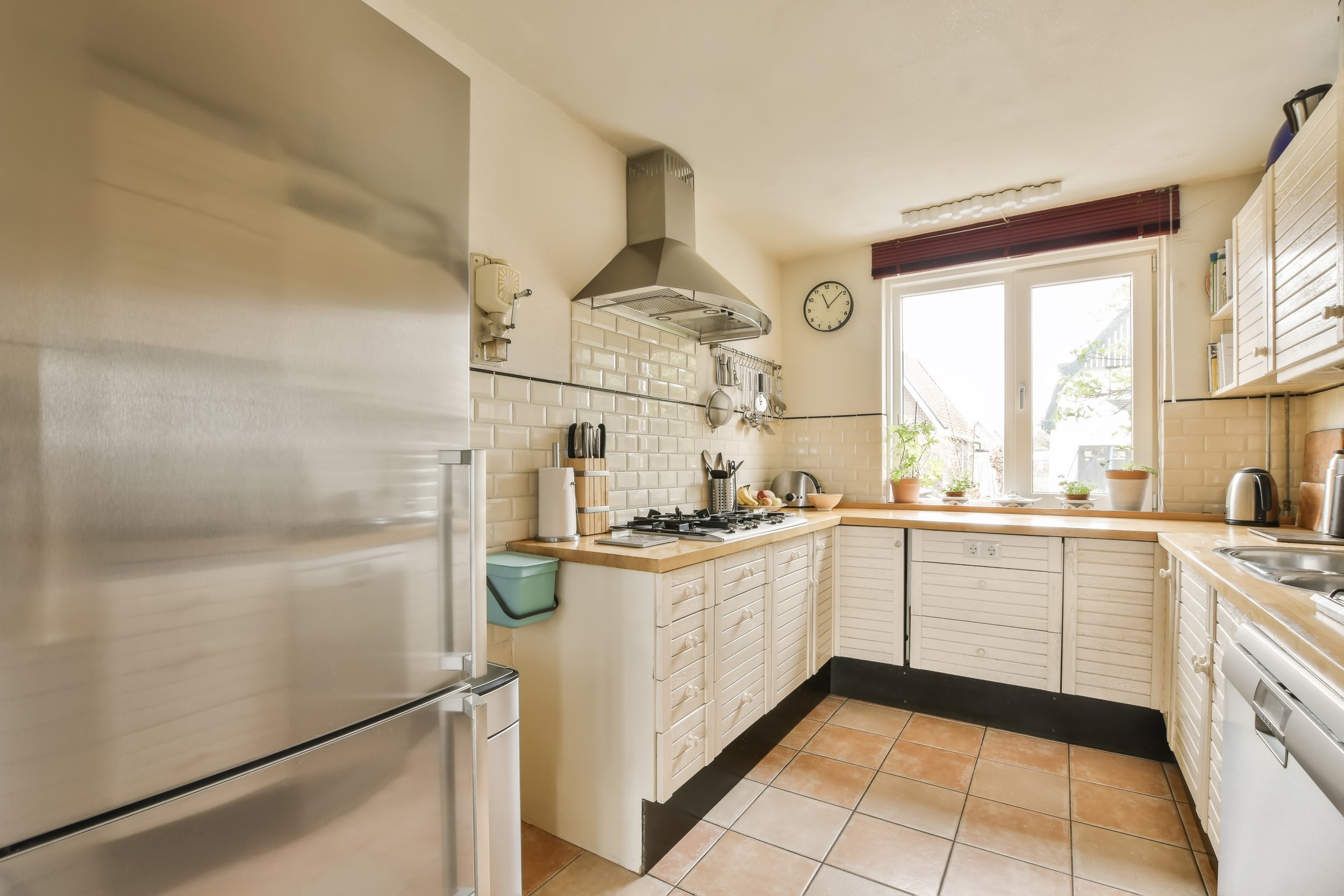 A kitchen with white cabinets, a window with a red blind, a stainless steel refrigerator, a gas stove with a range hood, and various kitchen utensils and plants on the countertop.