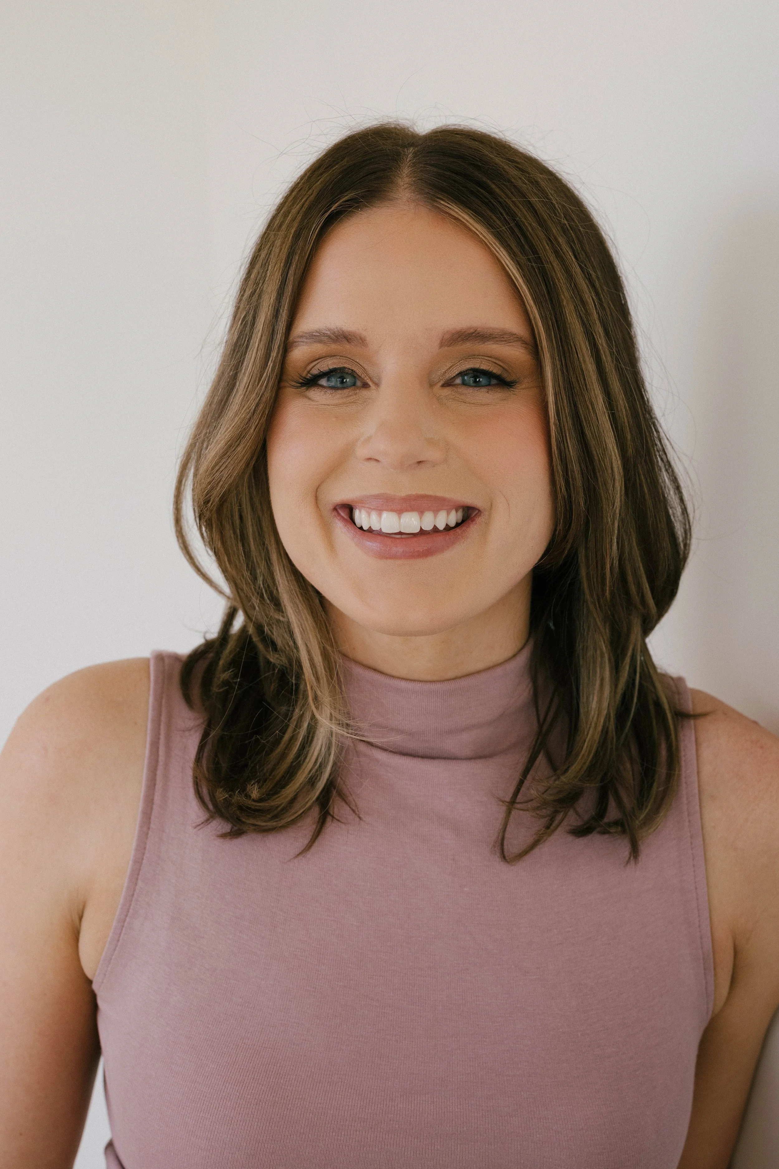 Close-up of a young woman with shoulder-length brown hair, wearing a sleeveless pink top and smiling against a plain light background.