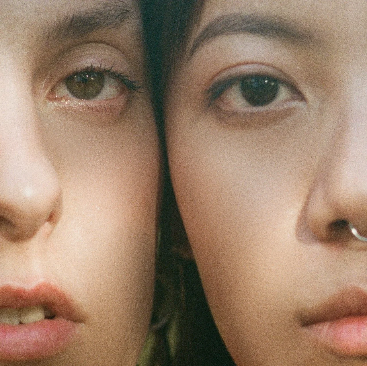 Close-up image of two faces, one with fair skin and the other with slightly darker skin, both with visible makeup and piercing eyes, positioned very close together.