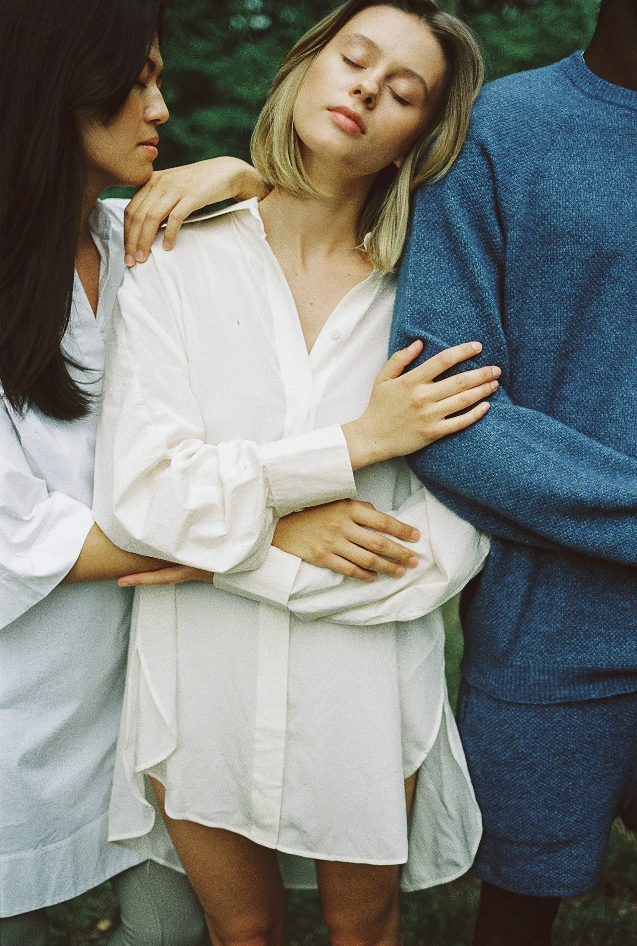 Three people standing closely together outdoors, with one woman resting her head on the shoulder of a person in a blue sweater. The woman in the middle has blonde hair and wears a white shirt. The person on the left has long dark hair, and the person on the right has dark skin.