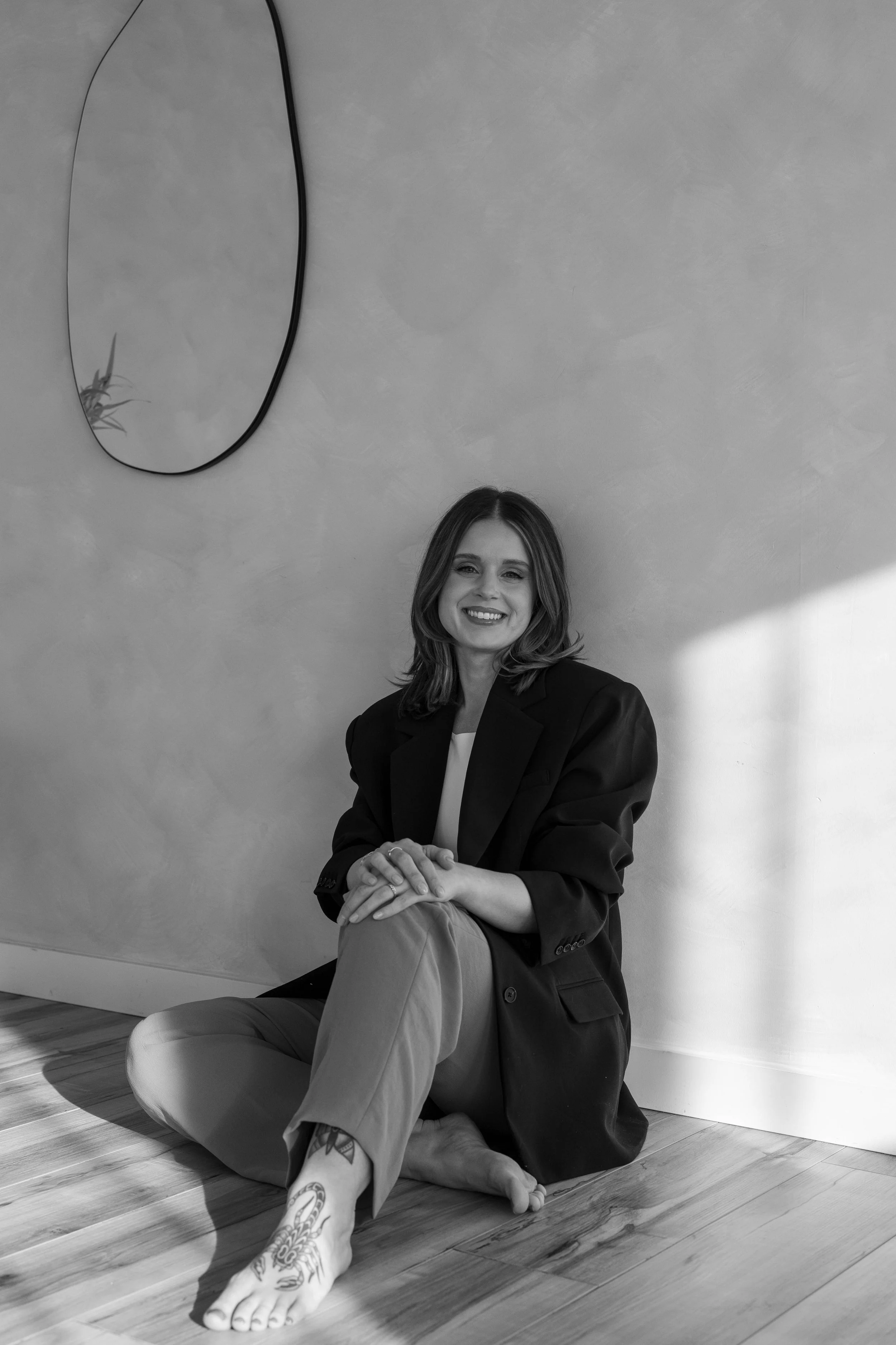 Black and white photo of a woman sitting on the floor with her legs crossed, smiling at the camera. She is dressed in a blazer and pants, with tattoos on her foot, and is in a room with a curved mirror on the wall and hardwood flooring.