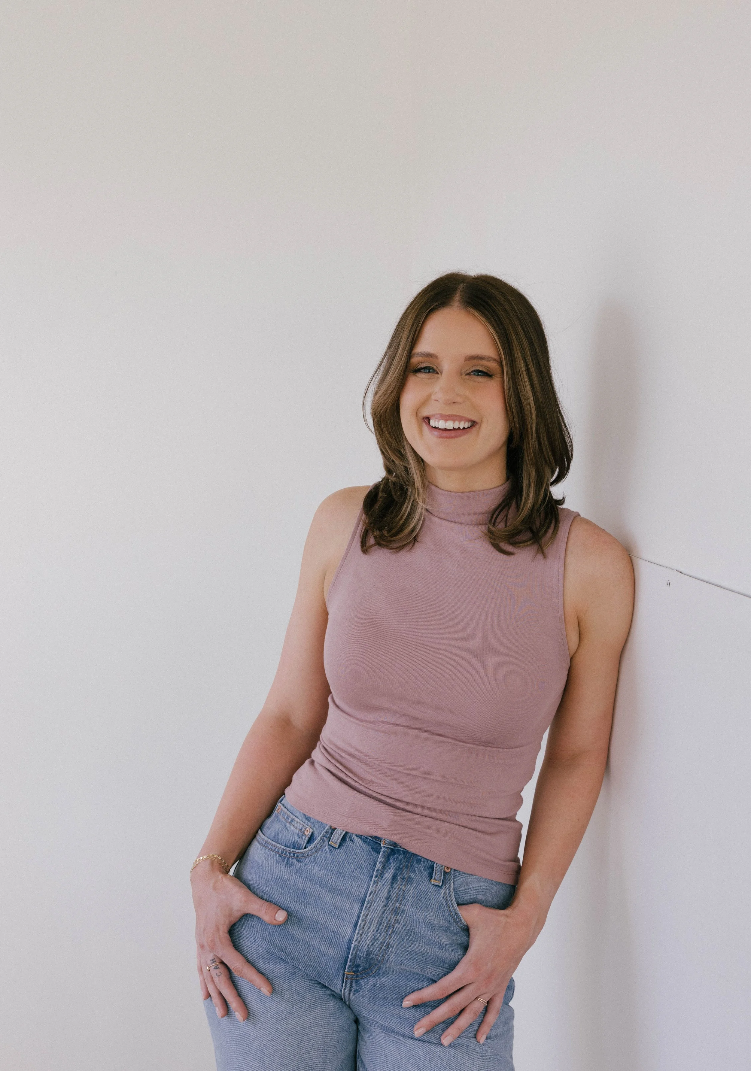 A young woman with shoulder-length brown hair is smiling and leaning against a white wall. She is wearing a sleeveless lavender top and light blue jeans, with her right hand resting on her hip and her left hand in her pocket.