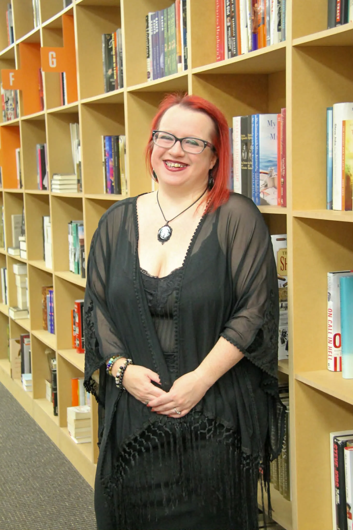 A woman with red hair and glasses, smiling, standing in a library in front of wooden bookshelves filled with books.