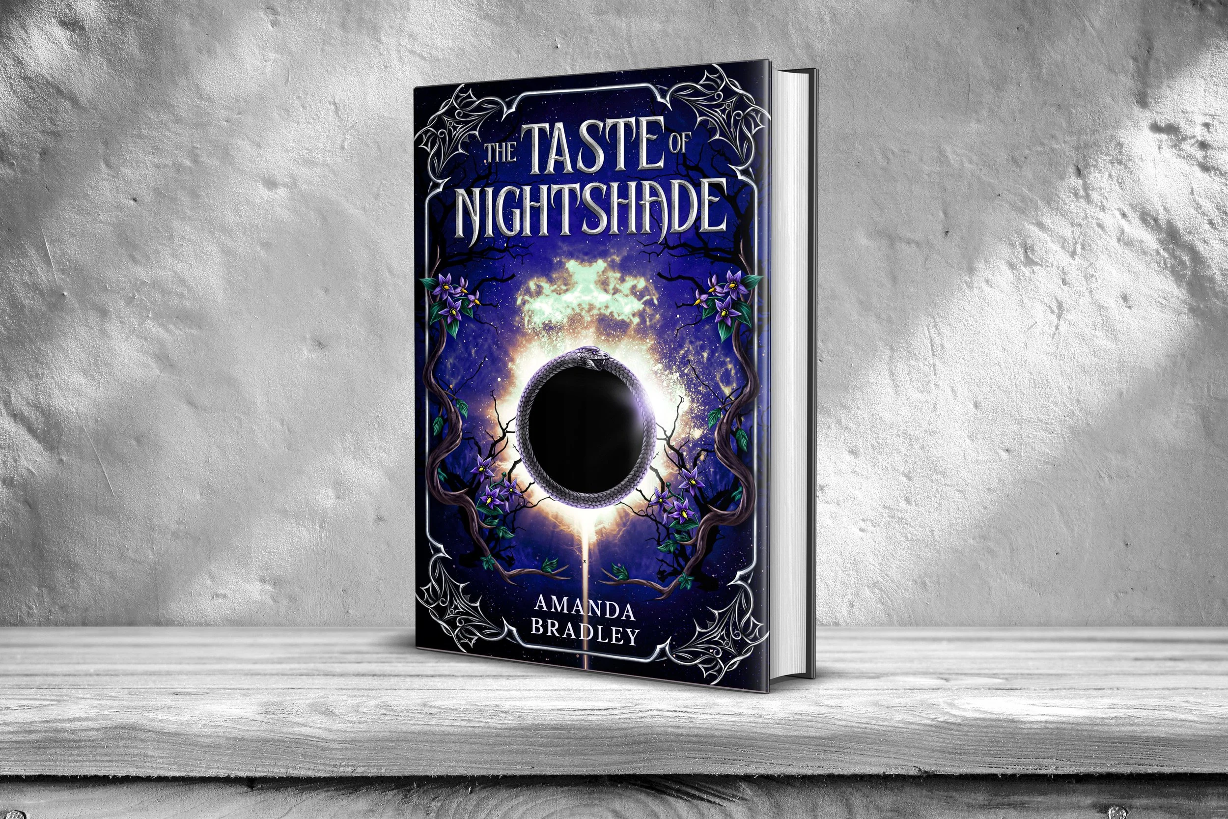 A hardcover book titled 'The Taste of Nightshade' by Amanda Bradley, featuring a dark, mystical cover design with a snake and purple flowers, placed on a wooden surface against a gray textured background.
