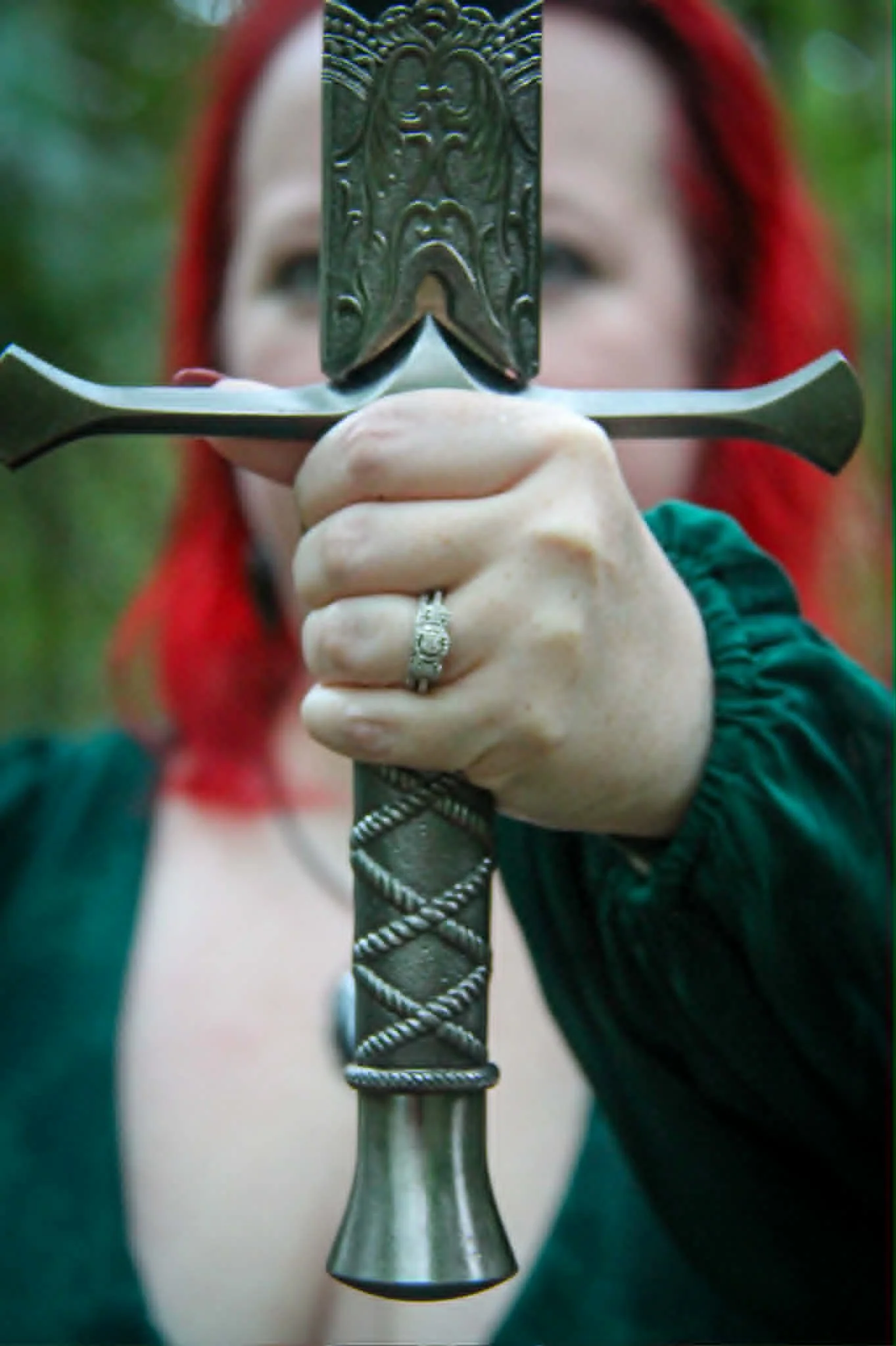 Person with red hair holding a sword vertically, with the blade close to the camera. The person's face is blurred and partially obscured by the sword. They are wearing a ring on their finger and a green jacket. The background is out of focus with green foliage.