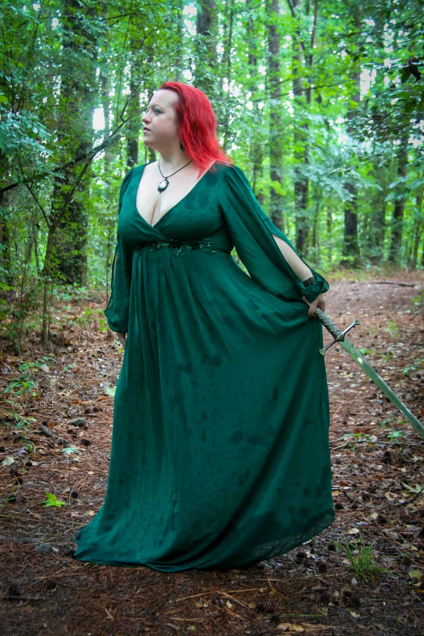 Woman with red hair in a long green dress standing in a forest holding a sword behind her back.