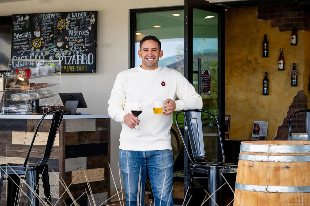 Man standing inside a bar or restaurant, holding a glass of red wine in one hand and a glass of beer in the other, smiling at the camera.