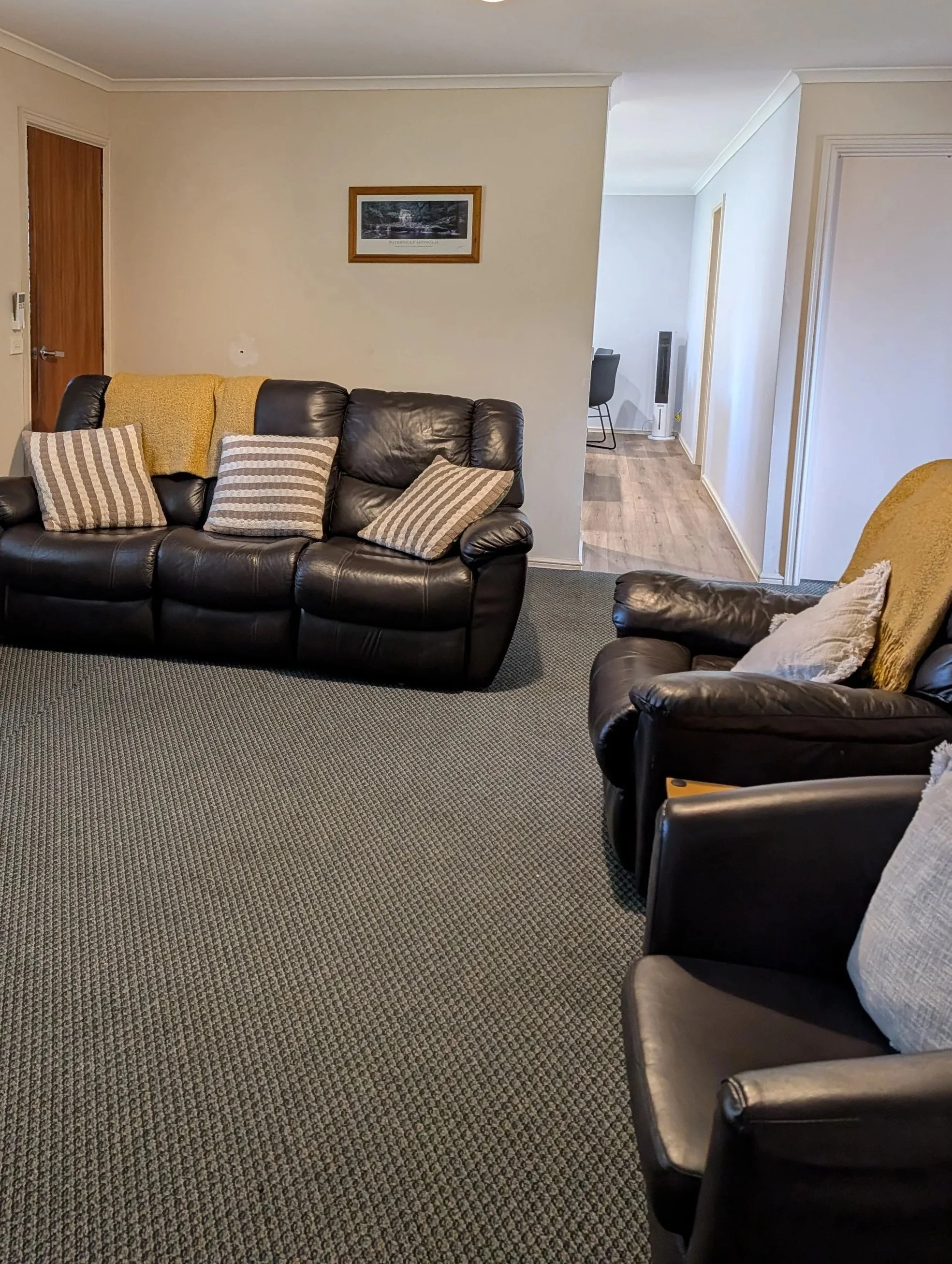 Living room with a dark brown leather couch, decorative pillows, yellow throws, a carpeted floor, and a hallway with wooden flooring leading to another room with a chair and a tower fan.