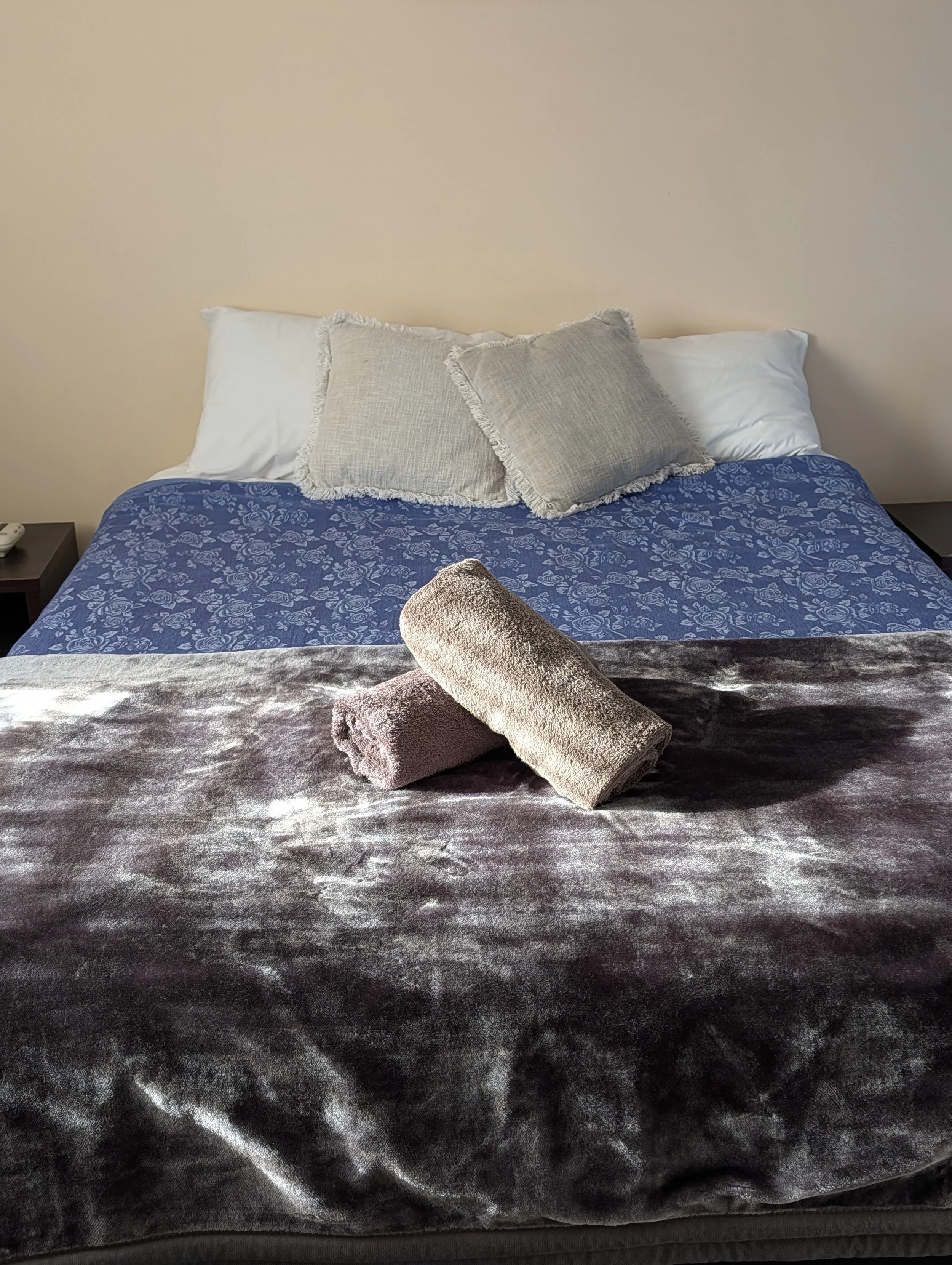 A neatly made bed with white pillows, beige throw pillows with fringed edges, a blue floral bedspread, and a rolled-up towel on top of a velvet blanket, with a beige wall in the background.