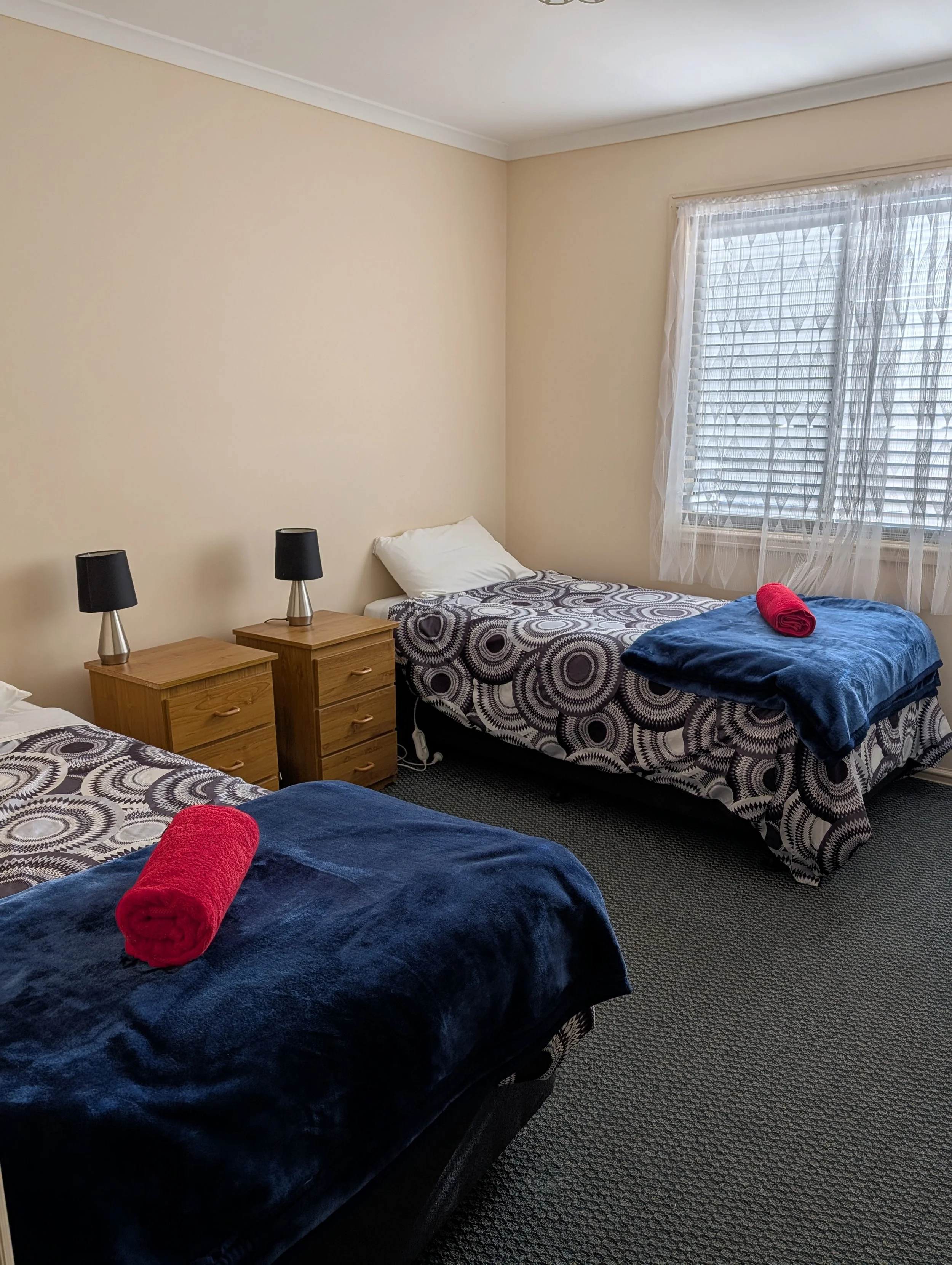 A bedroom with two beds, each with a patterned black and white bedspread, a rolled red towel on one bed and a rolled red towel on the other bed. Each bed has a pillow. Between the beds are two wooden nightstands, each with a black lamp. The room has beige walls, a window with white blinds, and sheer white curtains. The floor is carpeted in dark color.