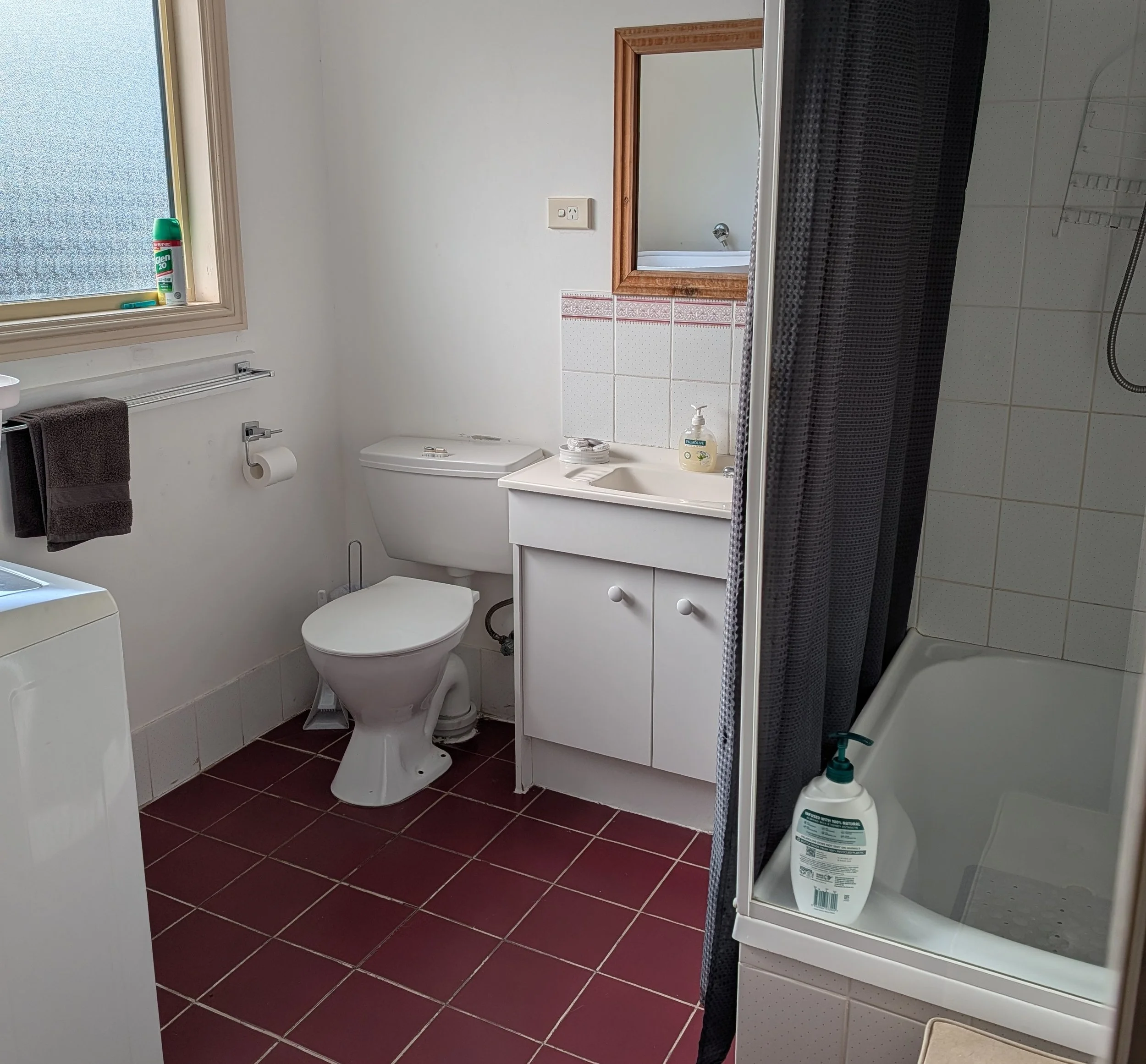 A small bathroom with white walls and maroon-tiled floor. There is a window with a frosted glass pane and a can of Glade air freshener on the window sill. The bathroom features a white toilet, a white sink with a soap dispenser and a mirror with a wooden frame above it. To the right, there is a shower with a dark striped curtain and showerhead. A bottle of cleaning product is on the floor next to the shower.