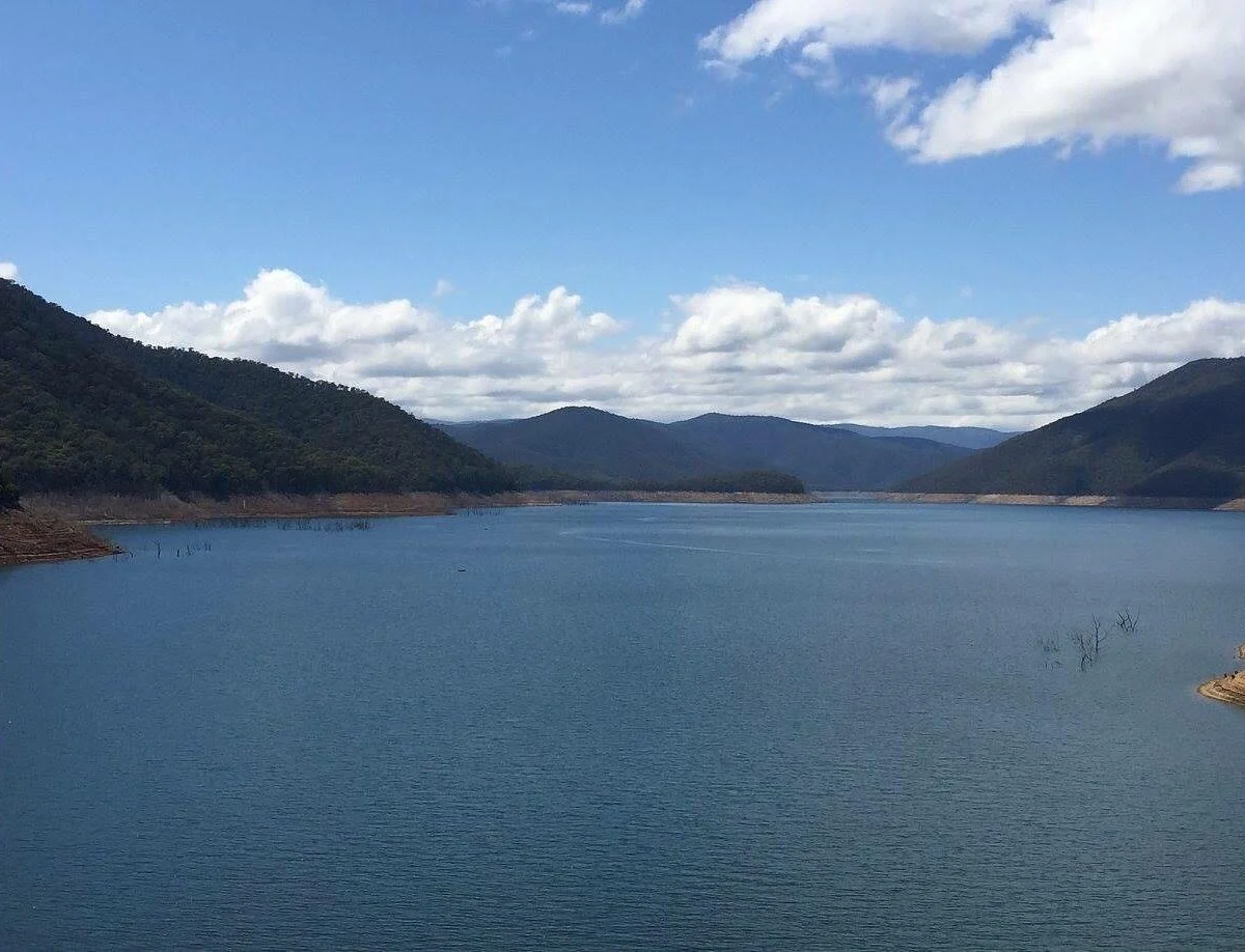 A large reservoir surrounded by green mountains under a partly cloudy blue sky.
