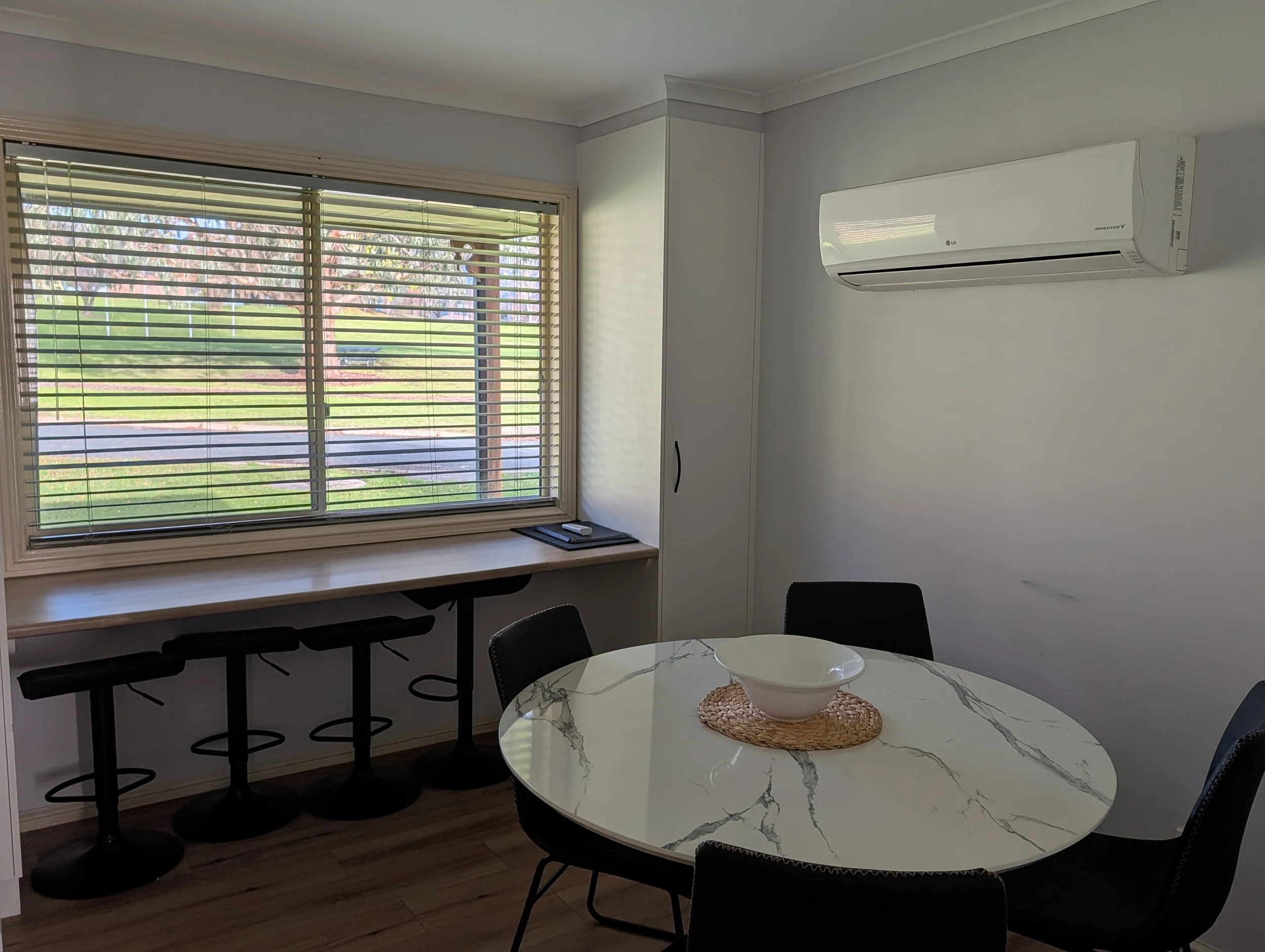 A room with a large window with closed blinds, an air conditioner mounted on a white wall, a white marble round dining table with black chairs, and a bowl on a woven placemat.