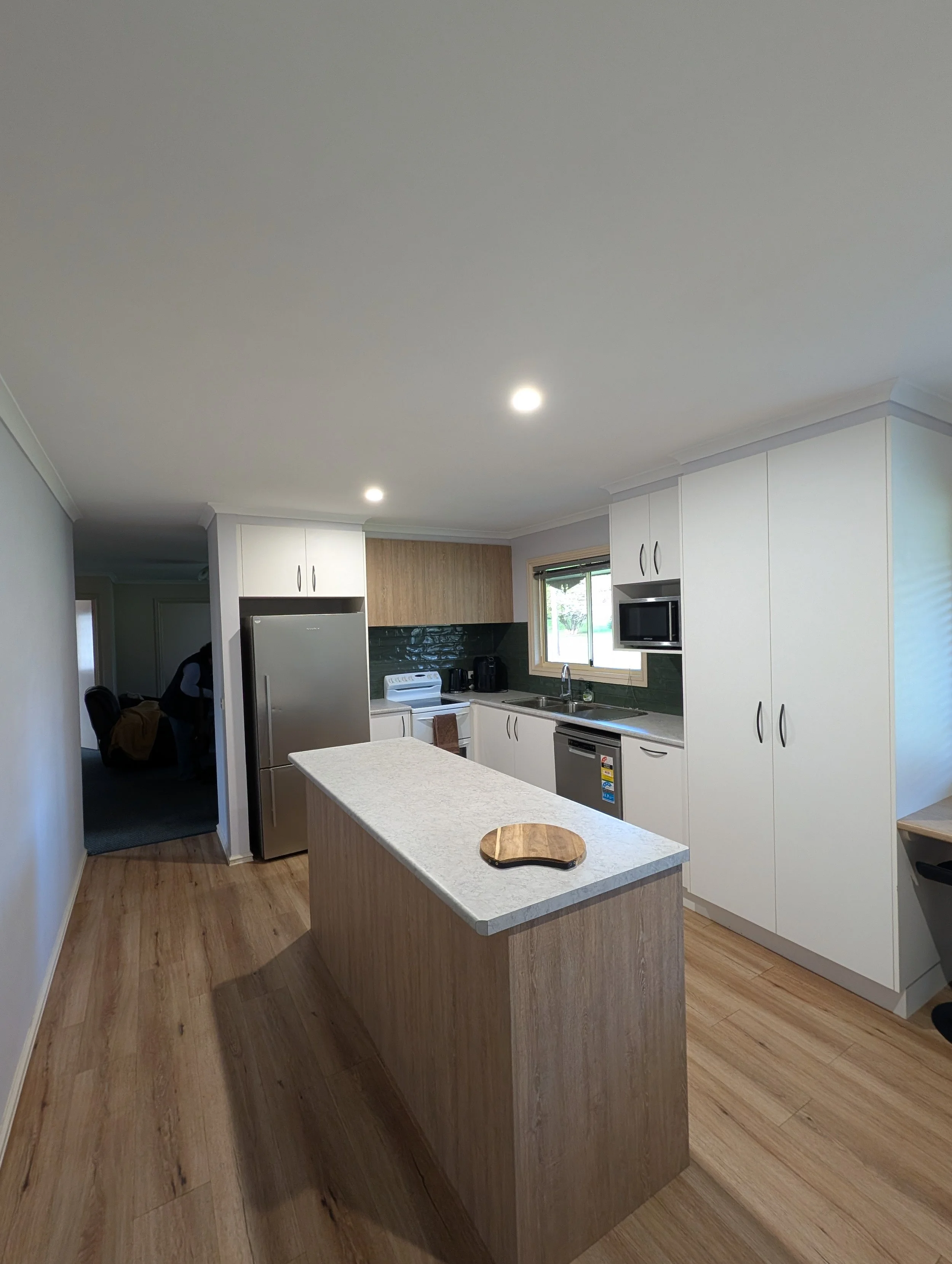 Kitchen with white cabinetry, a gray countertop kitchen island, a stainless steel refrigerator, microwave, oven, and window with a green view. Wooden flooring, ceiling lights, and a small wooden cutting board on the island.