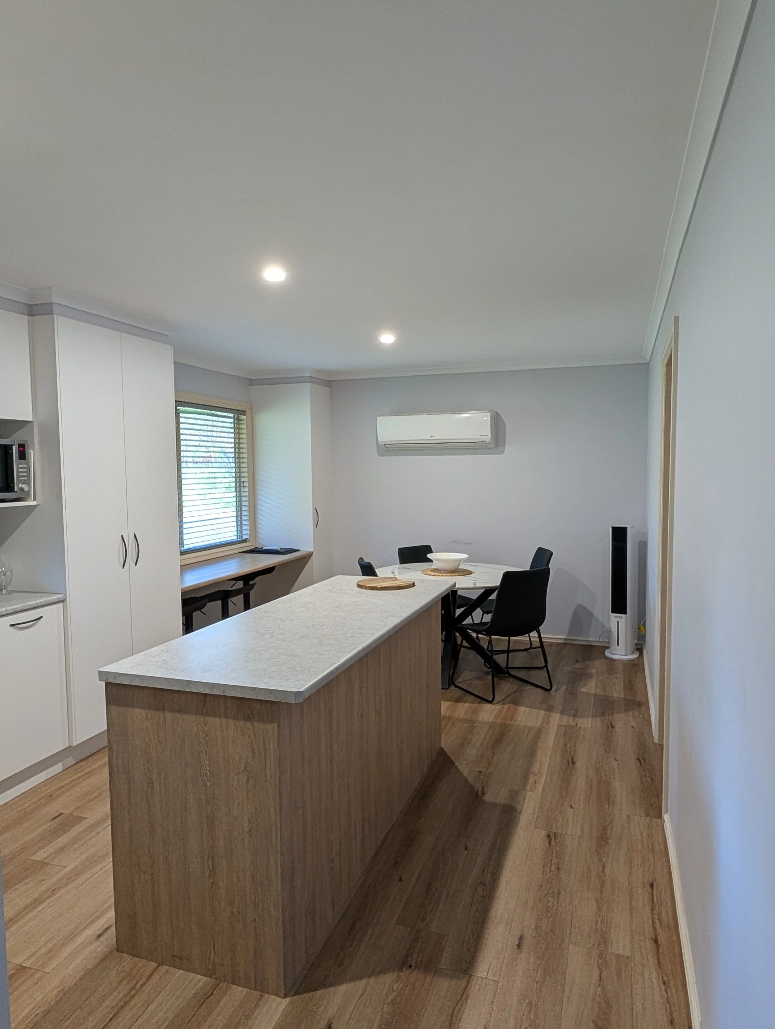 A modern kitchen with a white island, wooden flooring, white cabinets, a window with blinds, an air conditioning unit, and a dining table with chairs.