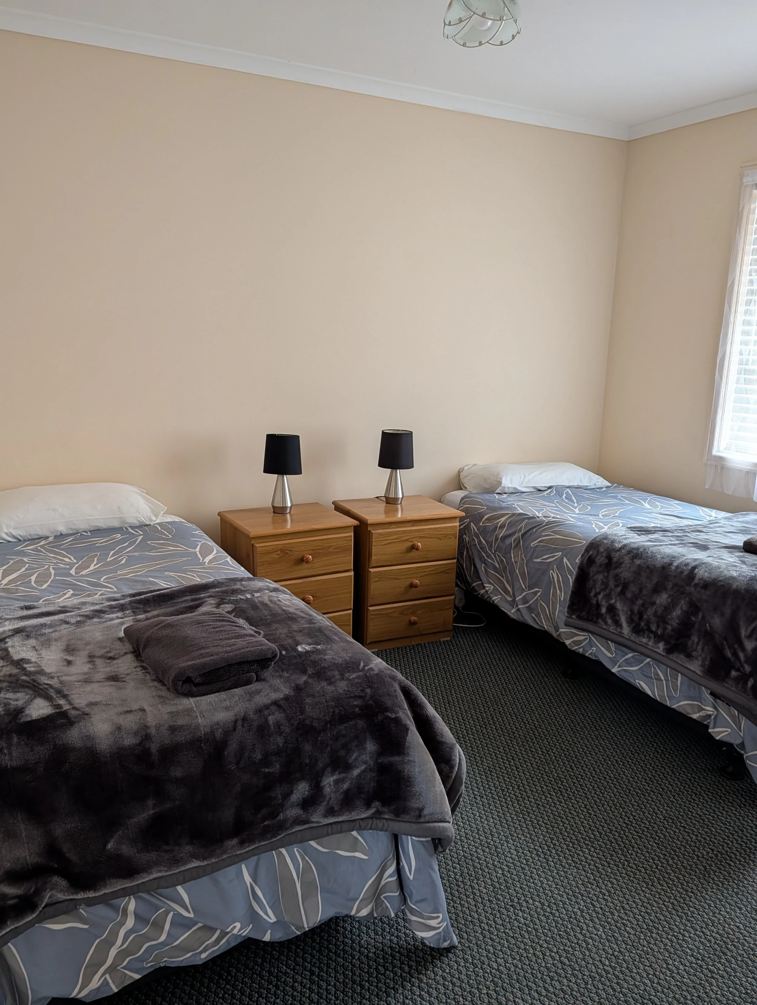 A bedroom with two beds, each with gray and white bedding, and a small wooden nightstand between them. On the nightstand are two black lamps. The room has a beige wall, a window with blinds on the right, and a ceiling light fixture.